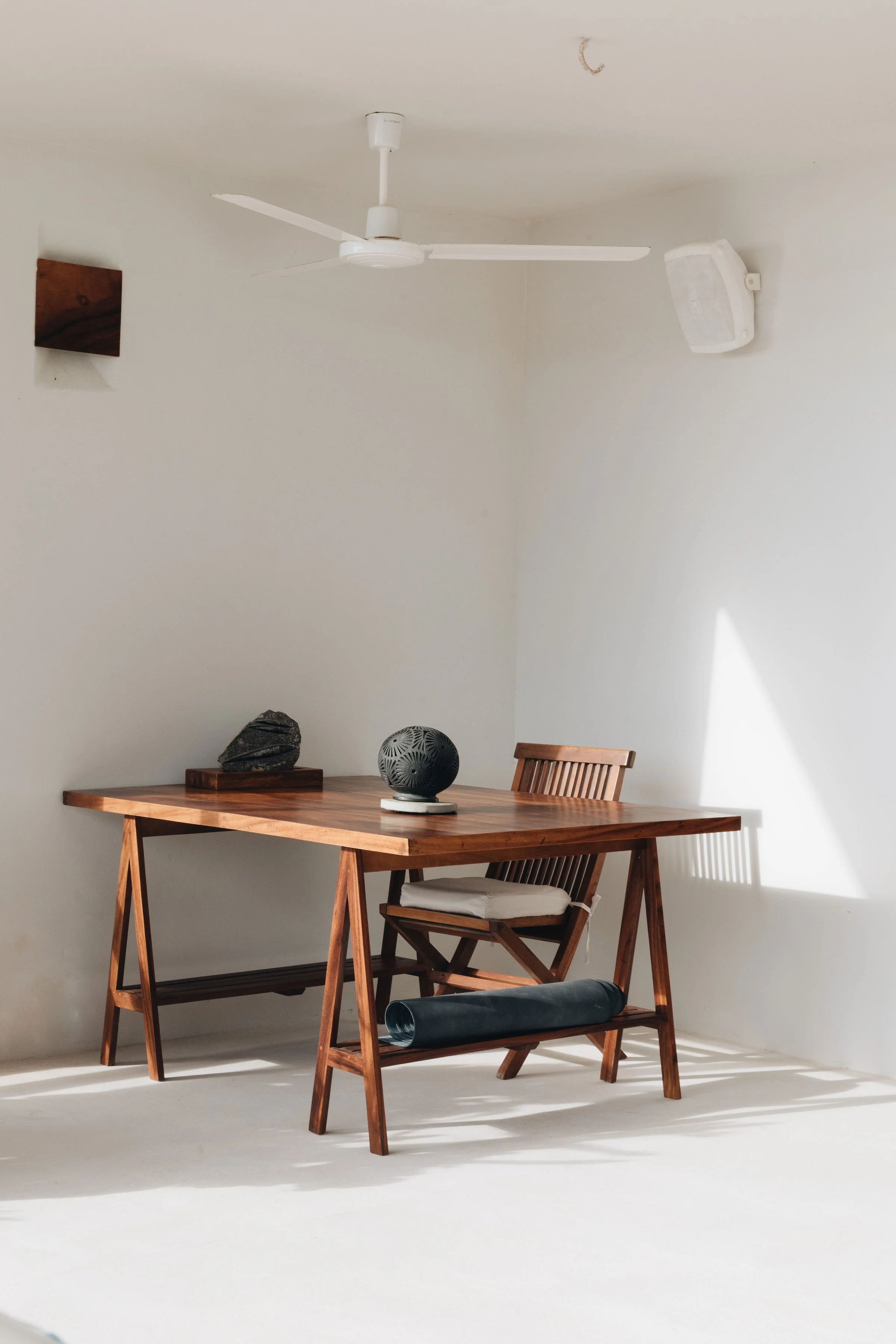 Mesa de madera con una silla, una escultura de piedra, una bola decorativa y un cilindro de material en la parte inferior, en una habitación con pared blanca y techo con ventilador y altavoz.