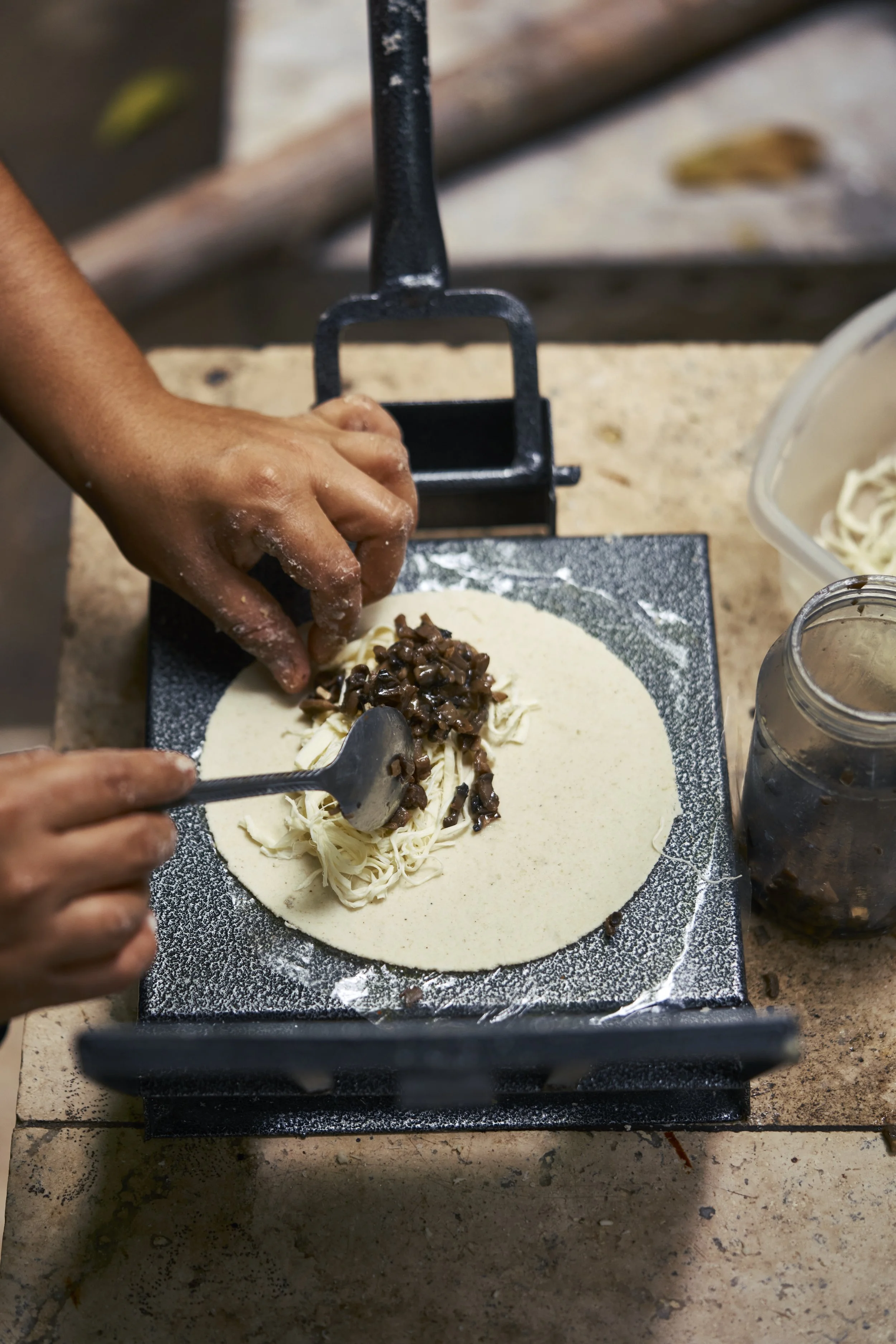 Persona colocando relleno de frijoles en una tortilla de maíz sobre una prensa para hacer tortillas.