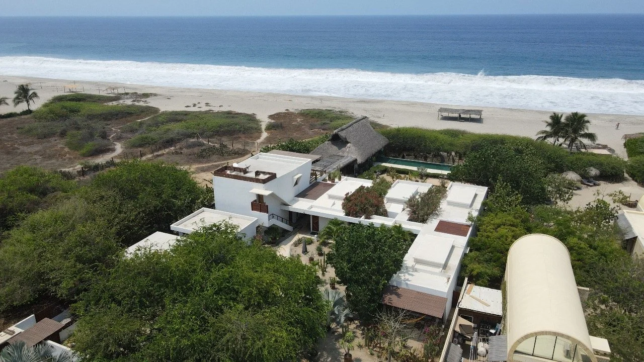 Vista aérea de una playa con olas y arena, y en primer plano, casas modernas y áreas verdes.