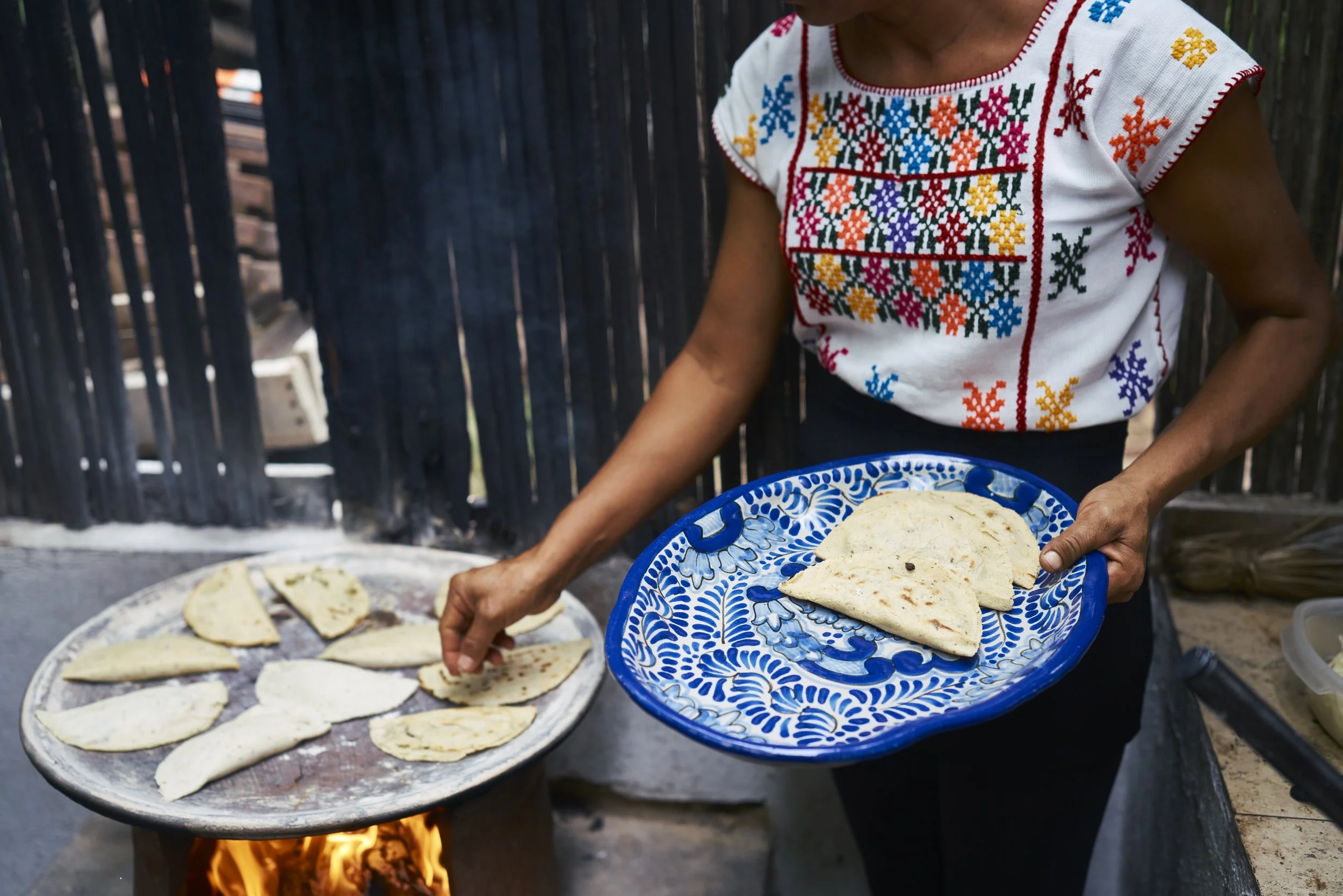 Una mujer vestida con una blusa bordada tradicionalmente, sosteniendo un plato azul con tortillas de maíz, mientras cocina en una fogata al aire libre.