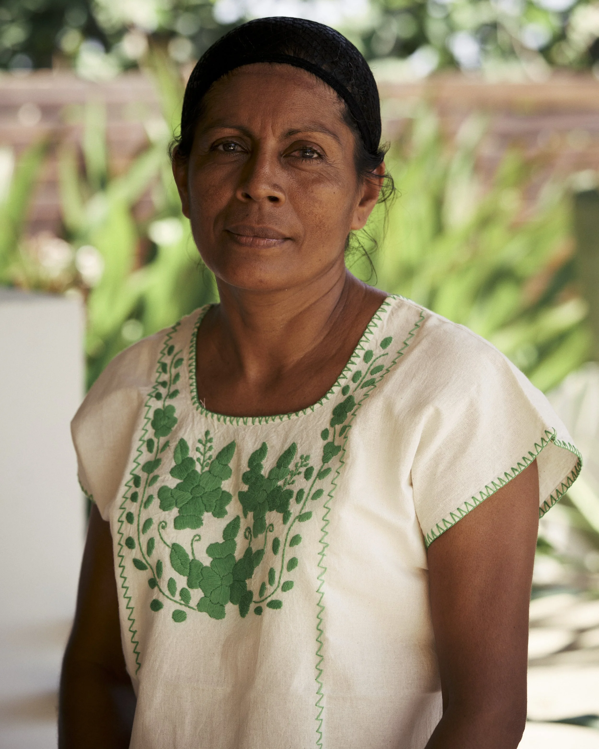 Mujer de piel morena, con cabello oscuro y trenza, vestida con blusa blanca con bordados verdes, en un entorno natural con plantas verdes de fondo.