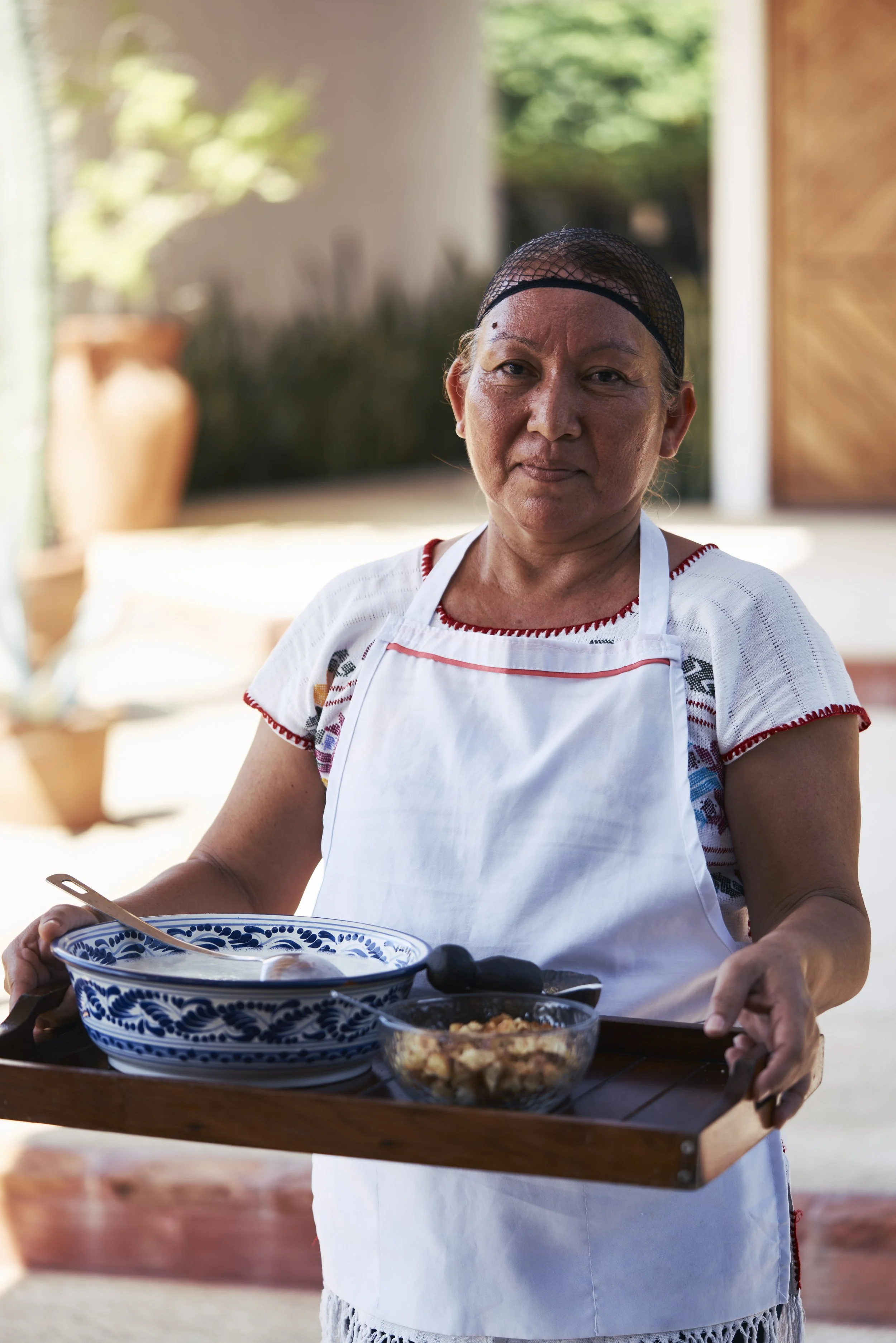 Mujer mayor con delantal blanco y gorra de malla negra sostiene una bandeja de madera con ingredientes de cocina, en fondo de casa y plantas verdes.