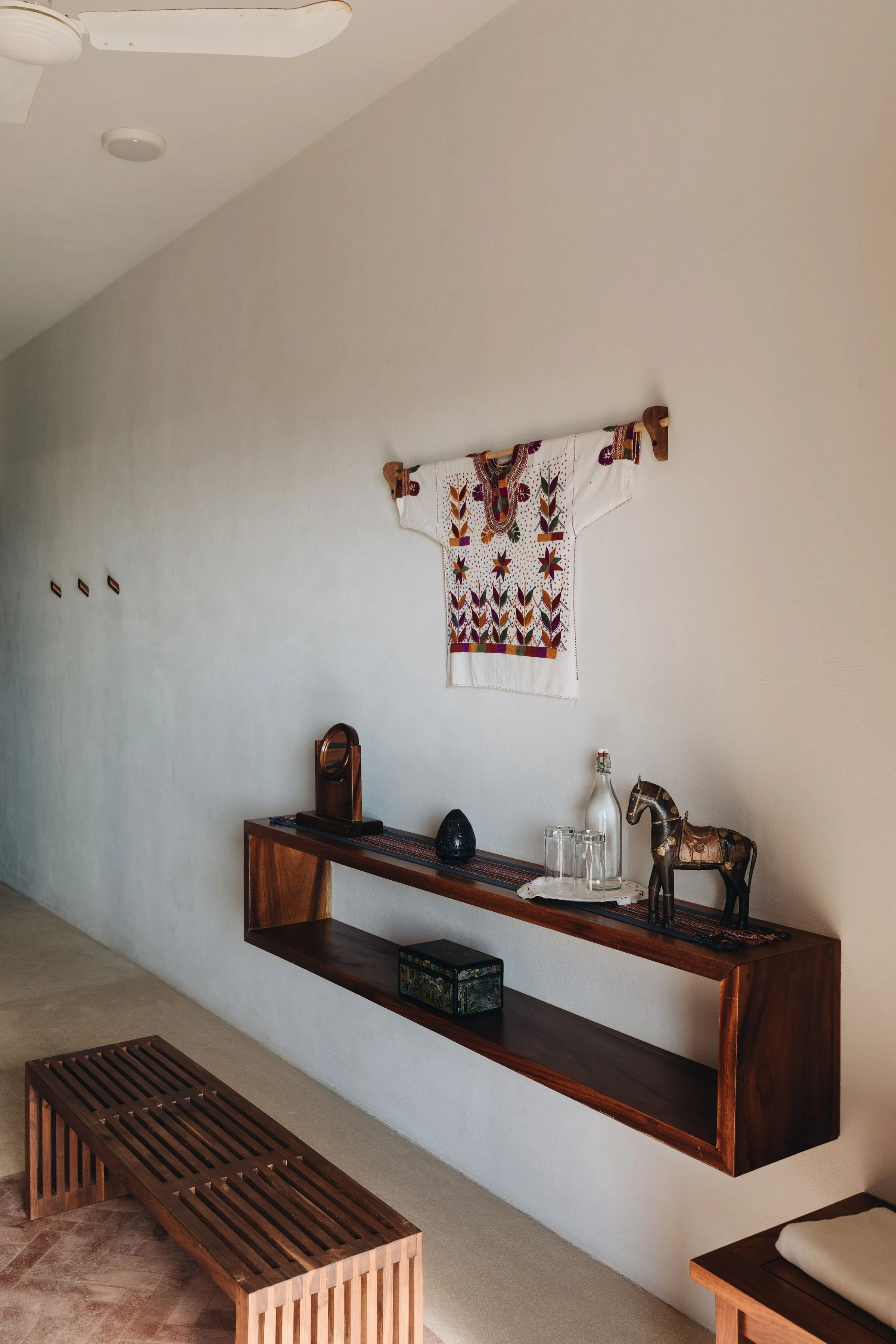 Decoración en la pared con un tapiz bordado, una repisa de madera con objetos decorativos y un taburete de madera.