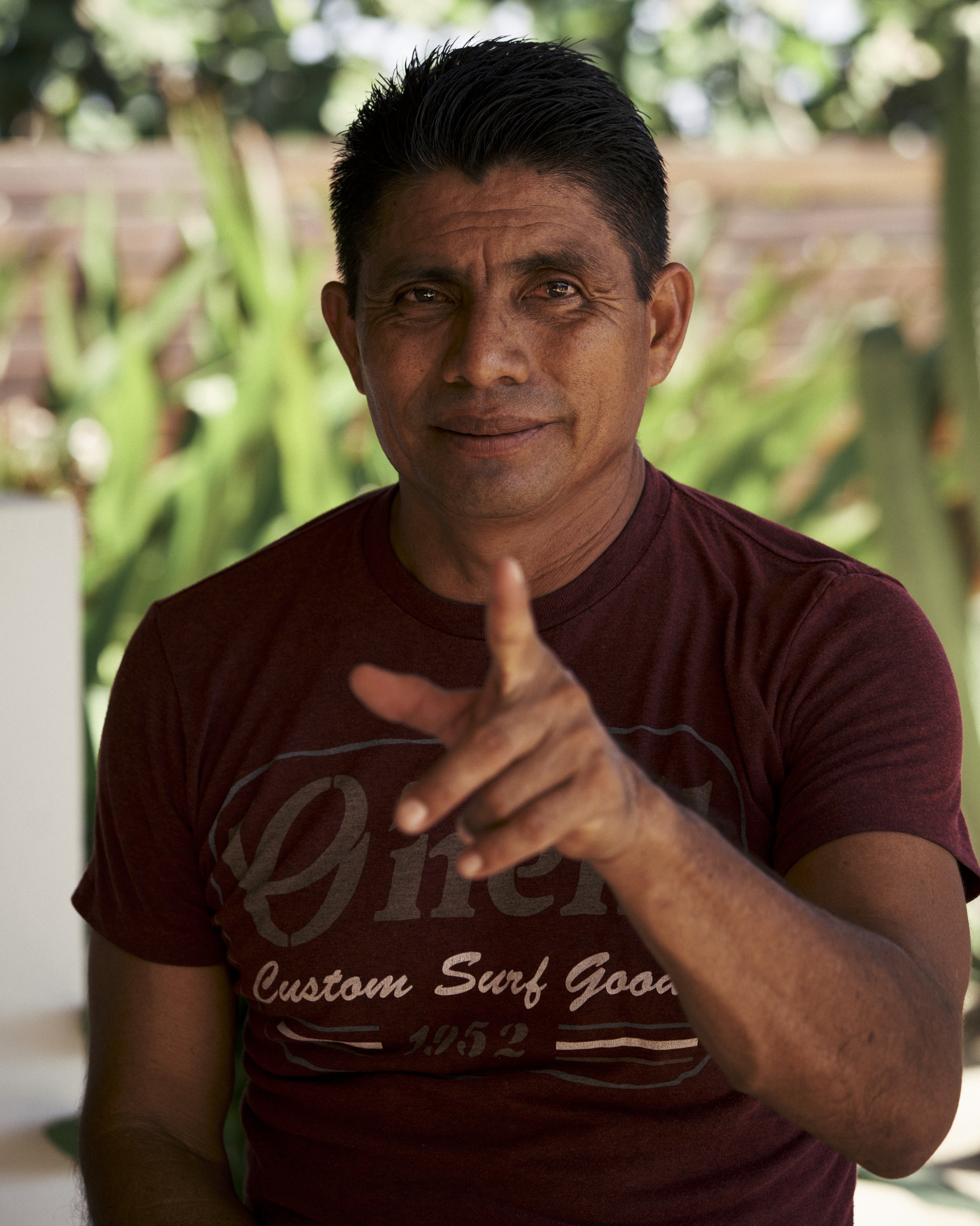 Hombre de piel morena, con cabello negro y peinado hacia arriba, viste una camiseta roja con letras y está señalando con el dedo índice hacia la cámara, en un fondo de vegetación.