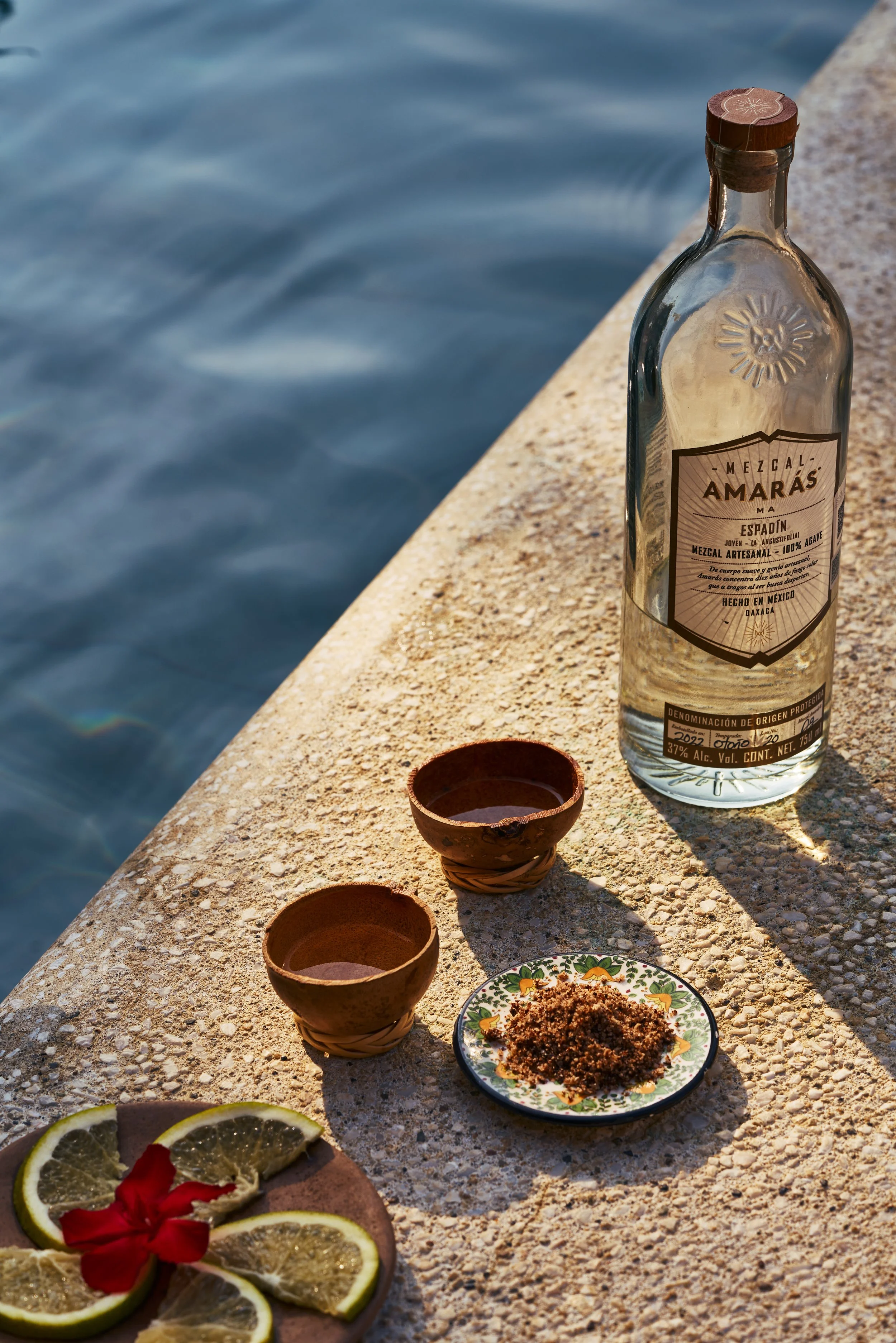 Botellas de mezcal con dos vasos pequeños y un plato con condimentos, junto a rodajas de limón y una flor roja al lado, en una superficie cerca del agua.