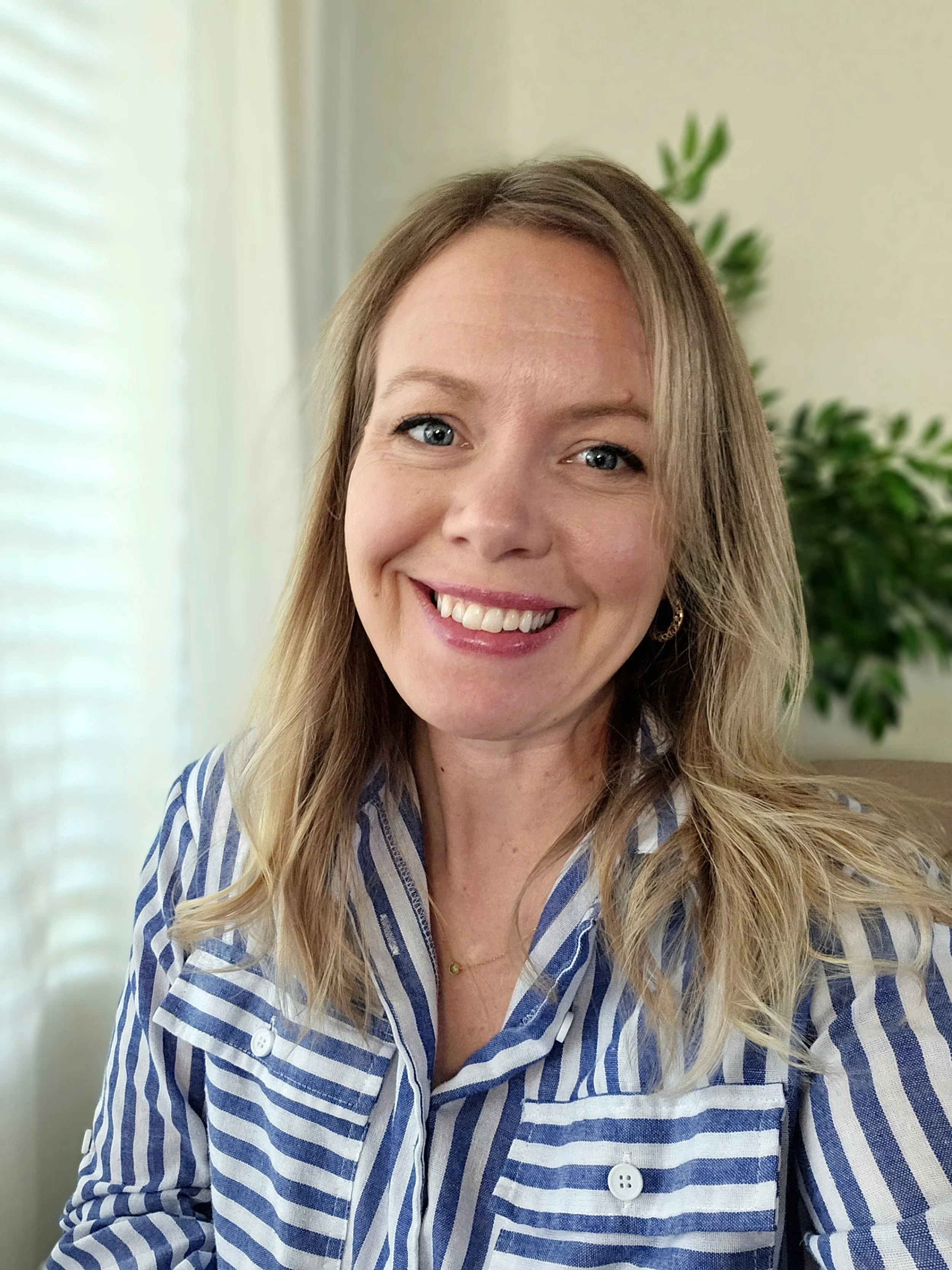 A woman with blonde hair, smiling, wearing a blue and white striped shirt, sitting indoors near a window with blinds and a green plant in the background.