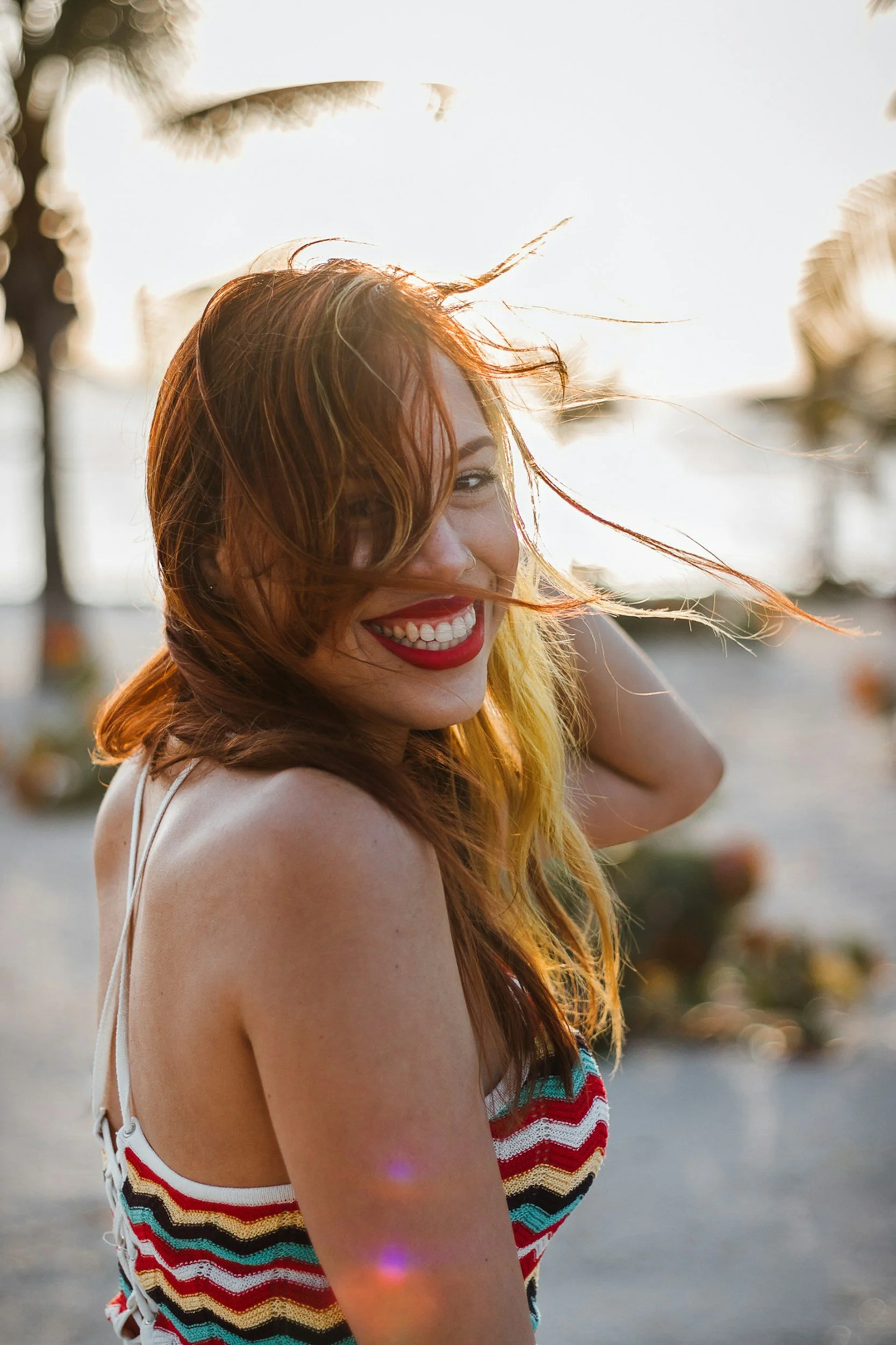 A young woman with red hair and red lipstick smiling on a beach during sunset, wearing a colorful striped dress.