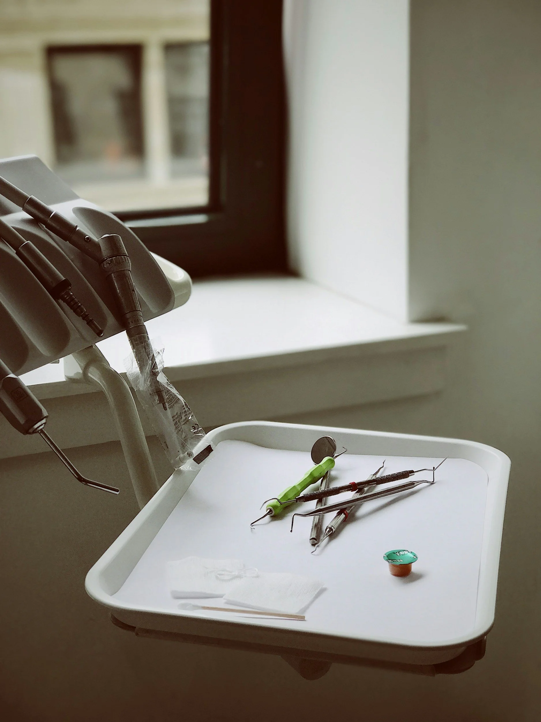 Dental tools on a tray including dental probes, mirror, and other instruments, near a dental chair and window.