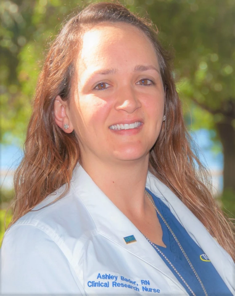 A woman with long wavy hair wearing a white medical coat and a blue shirt, standing outdoors with greenery in the background.