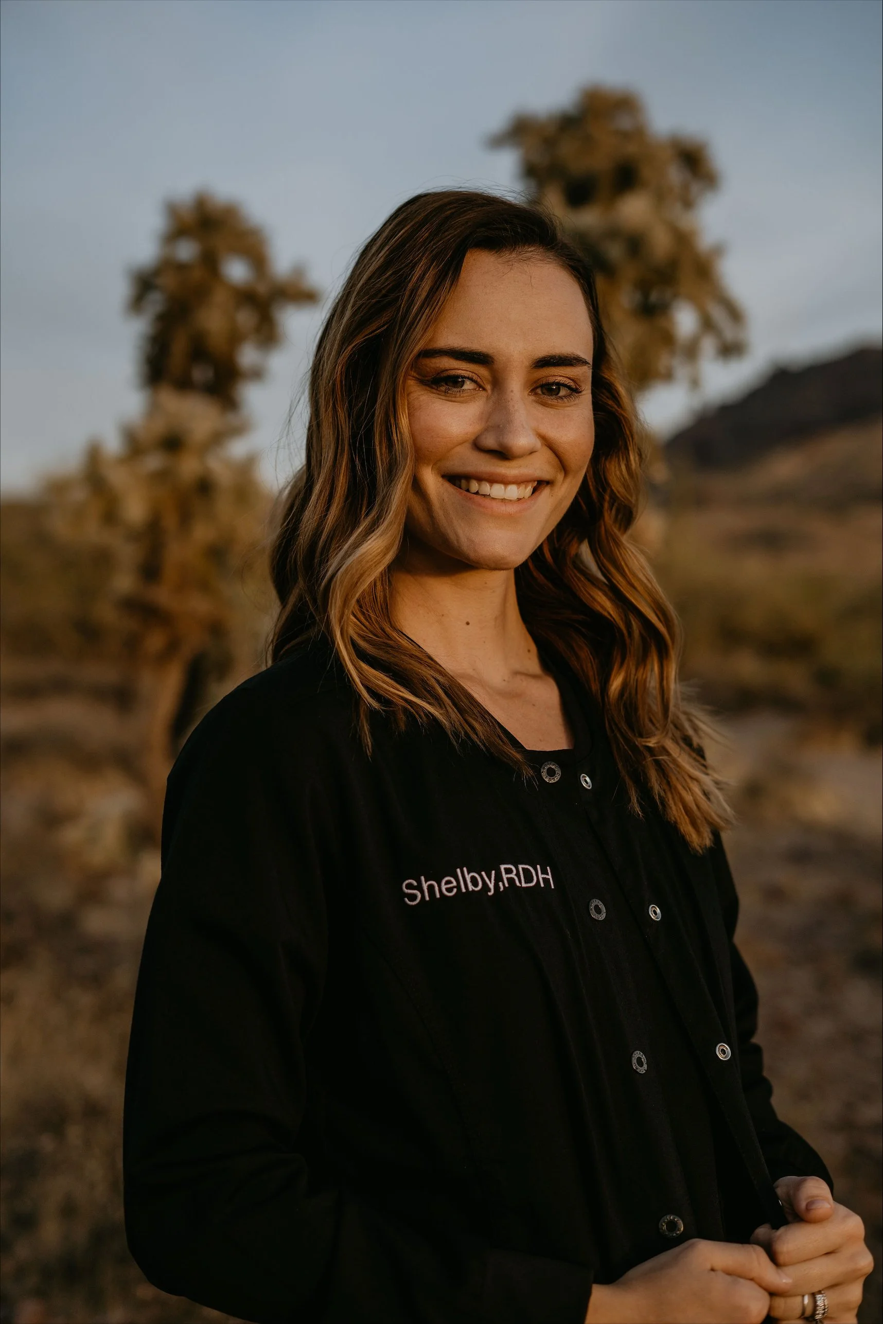A woman smiling outdoors during sunset, wearing a black jacket with 'Shelby, RDH' embroidered on it, standing in a desert landscape with Joshua trees in the background.