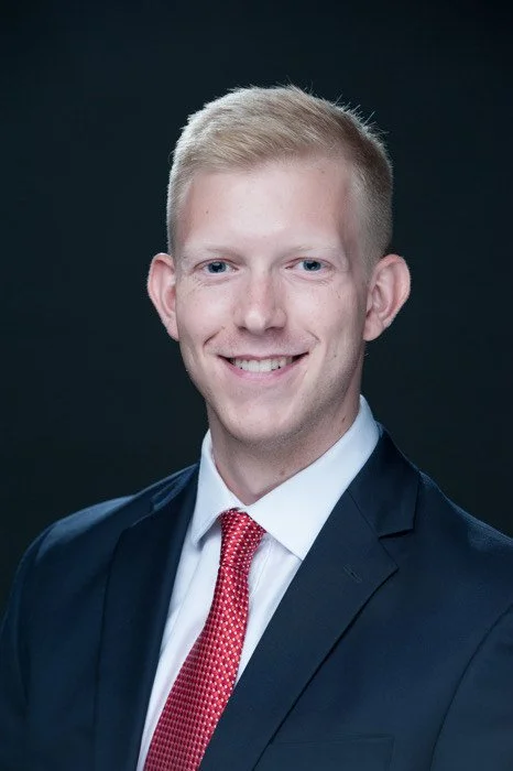 A young man in a dark suit with a white shirt and red tie, smiling, against a dark background.