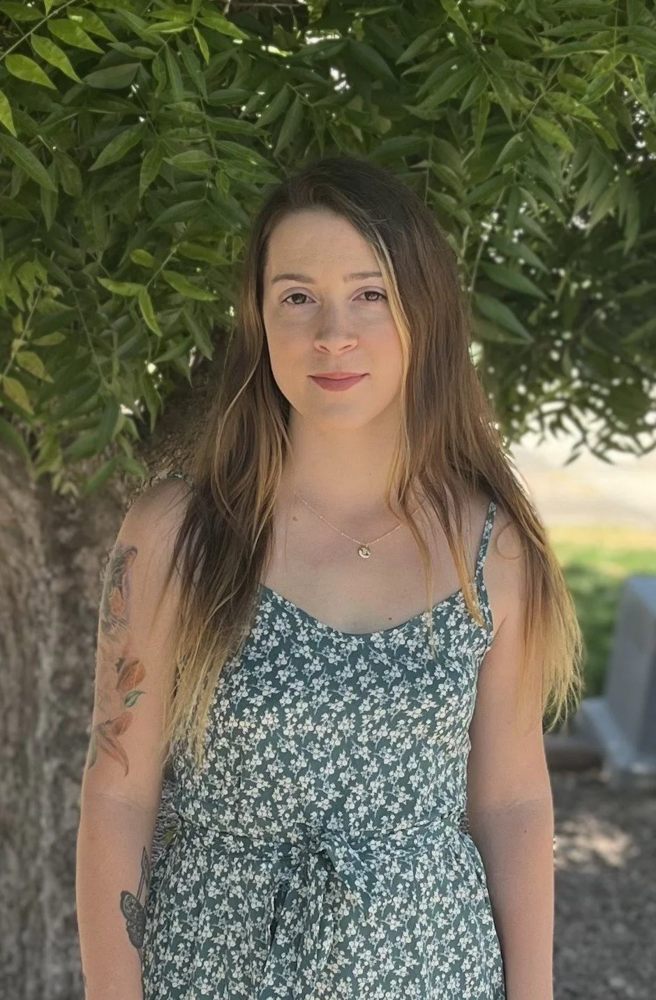 A young woman standing outside under a leafy tree, wearing a floral dress, with long brown hair, and tattoos on her left arm.