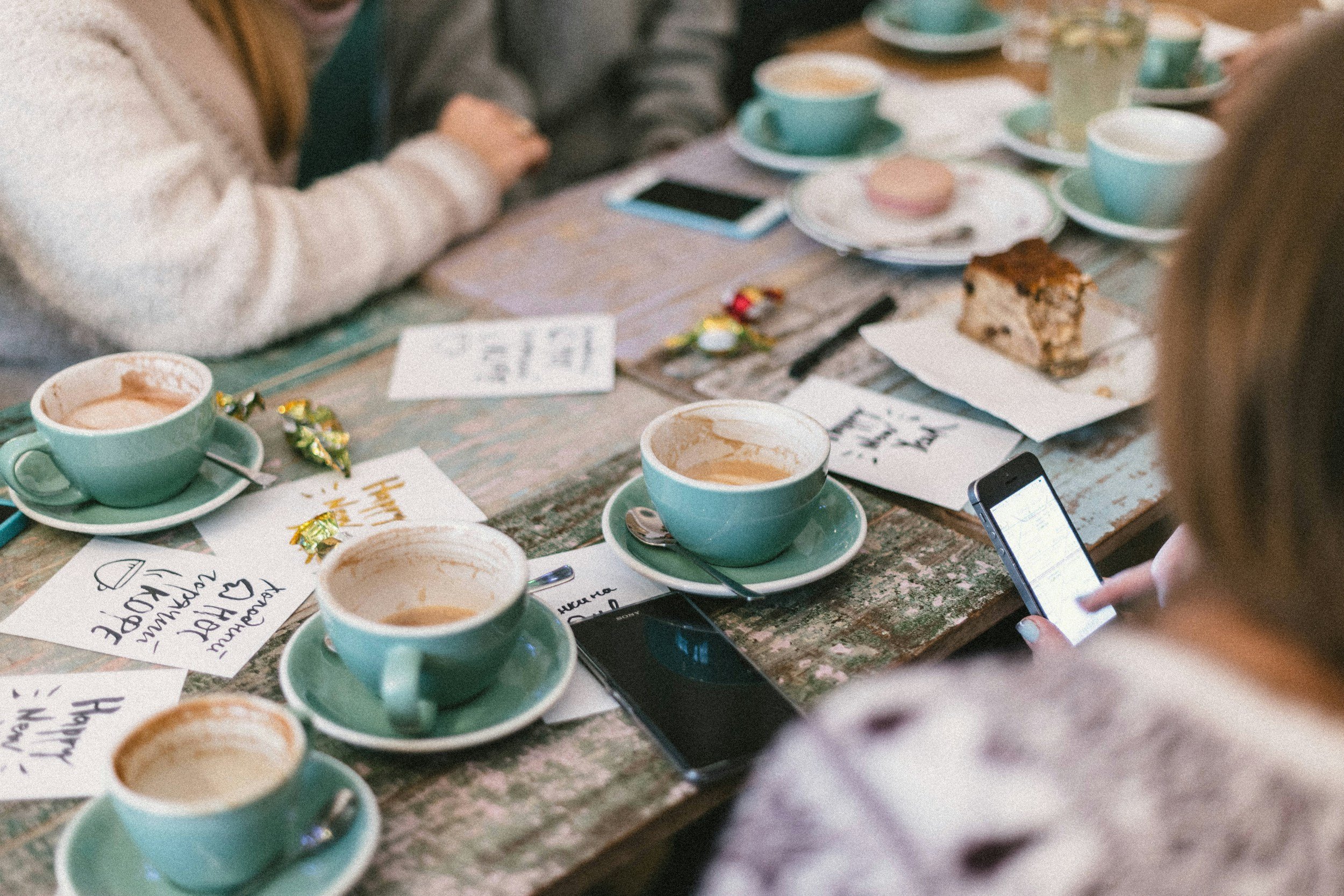 Groupe de personnes autour d'une table avec des tasses de café, des assiettes avec un gâteau, des cartes avec des messages, des téléphones portables et des bonbons. Le cadre semble être une réunion ou une célébration conviviale.