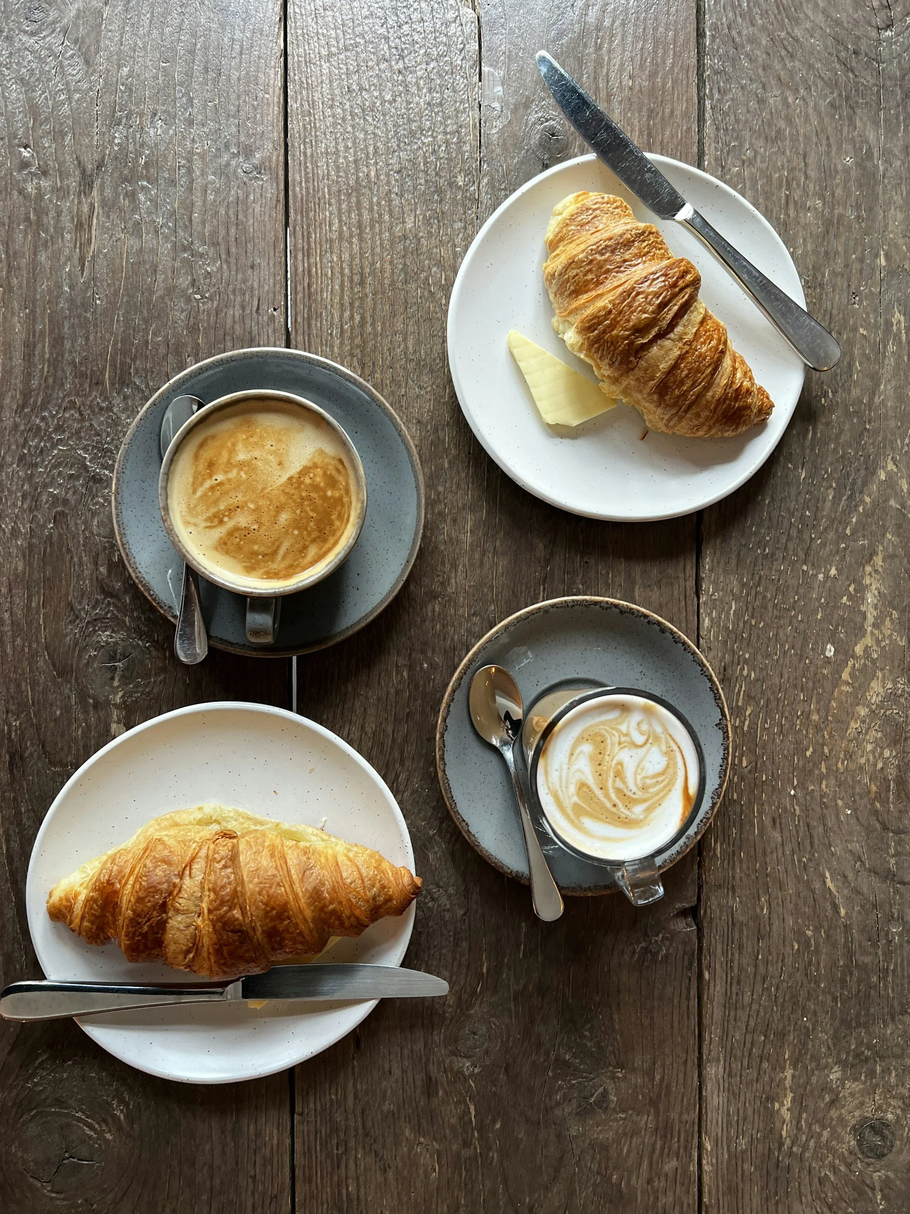Deux croissants sur des assiettes blanches, deux cafés avec crème sur des tasses grises, tous placés sur une table en bois