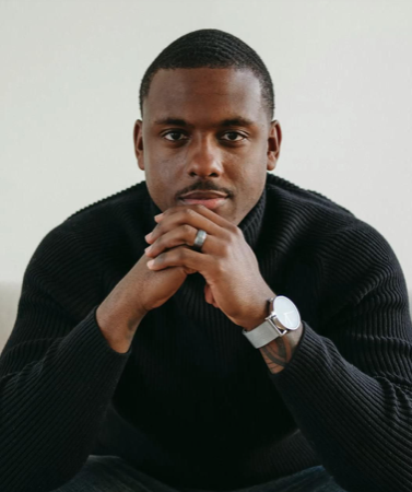 A young man with black hair smiling widely, surrounded by green plants indoors.