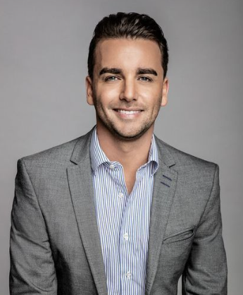 A man with dark, curly hair, clear glasses, and a beard and mustache, wearing a dark sweater. He appears to be sitting indoors near a plant with long, green leaves.
