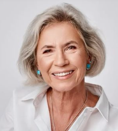 A young woman with curly hair, wearing glasses, earrings, and necklaces, smiling in a warmly lit indoor setting.