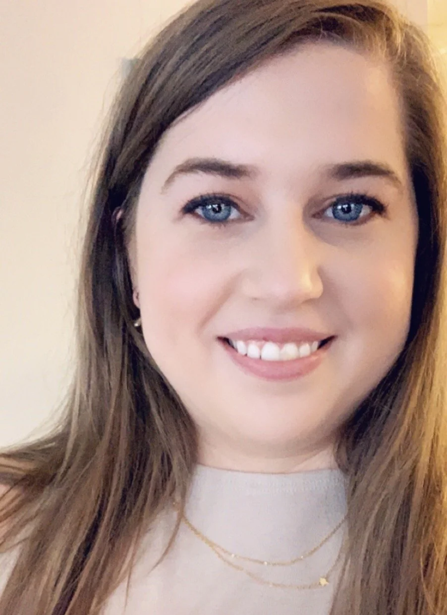 A woman with long brown hair, blue eyes, fair skin, smiling. She is wearing a light-colored top, small earrings, a gold necklace, and a delicate chain with a small star pendant.