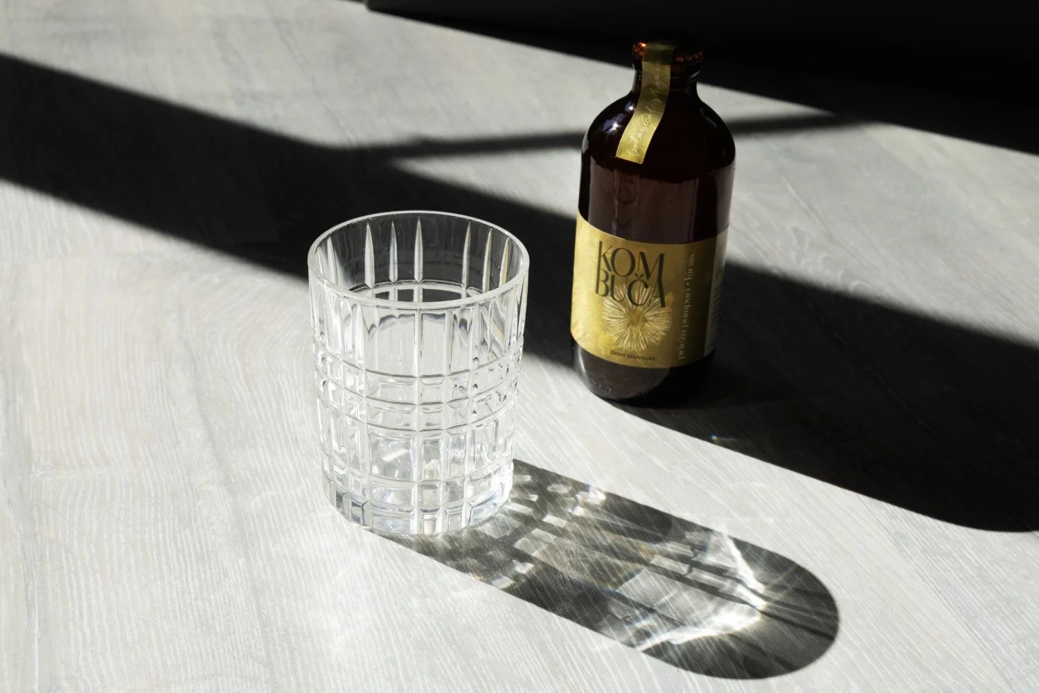 Kombuča pudele un stikla glāze dabīgā saules gaismā ar izteiktām ēnām