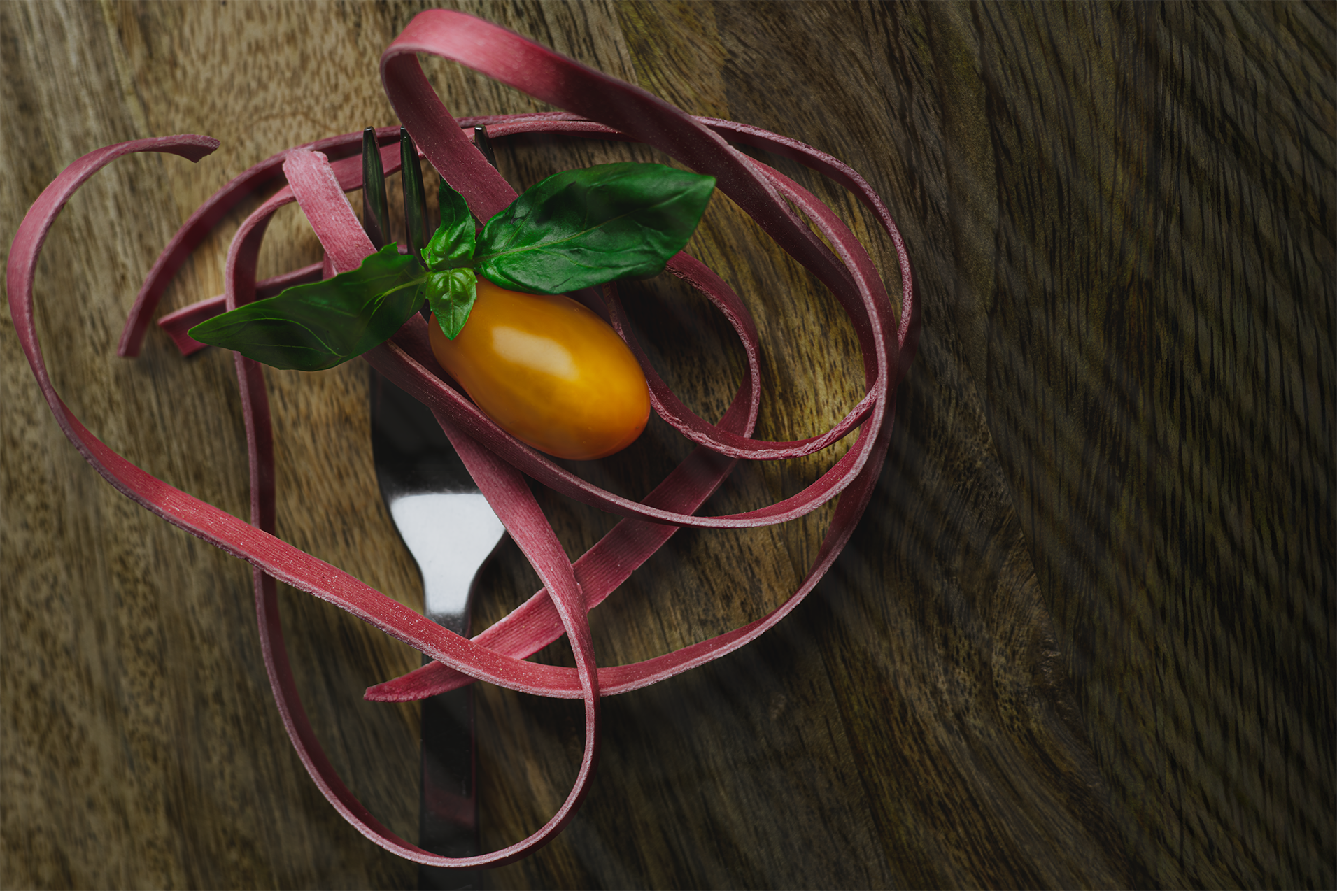 Mākslinieciski stilizēta violeta pasta ar bazilika lapām un dzeltenu tomātu uz tumšas koka virsmas.