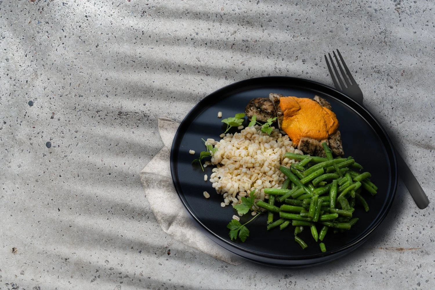 Minimālistiska ēdiena produktu fotogrāfija, kas uzņemta profesionālā studijas vidē. Uz tumšas, matētas šķīvja kompozīcijas redzama sabalansēta maltīte – grilēta gaļa ar krēmīgu mērci, pērļu grūbas un svaigi zaļās pupiņas. Fotogrāfija veidota no augša