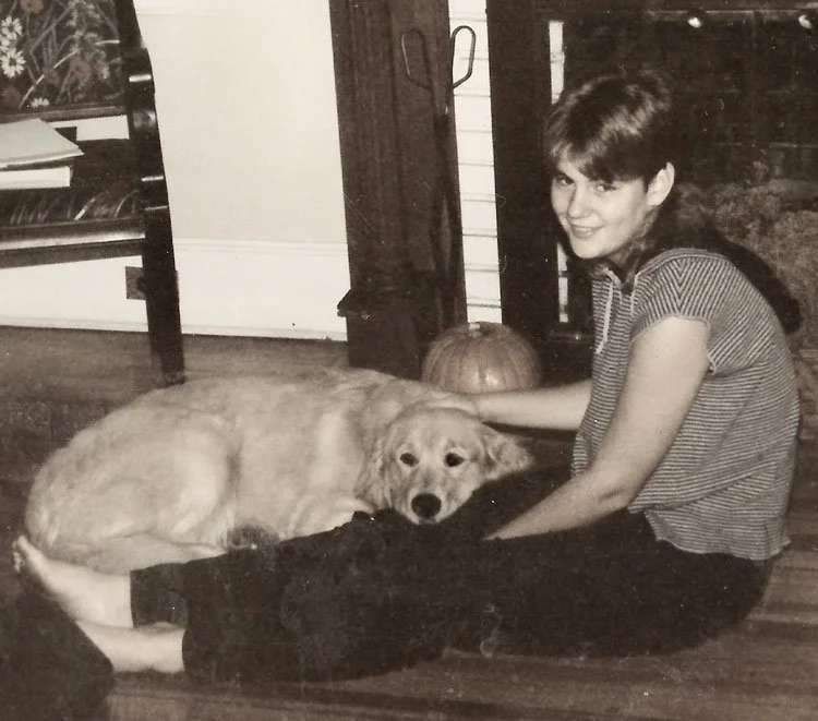 Owner at a young age sitting on the floor with her golden retriever dog lying beside her, both facing the camera, in a cozy living room with a pumpkin on the floor behind them.
