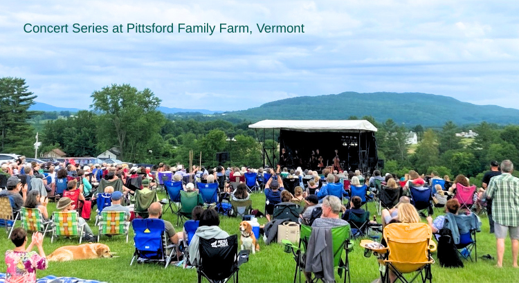 An outdoor concert at Pittsford Family Farm in Vermont, with a large crowd seated on the grass in front of a stage, surrounded by green trees and hills.