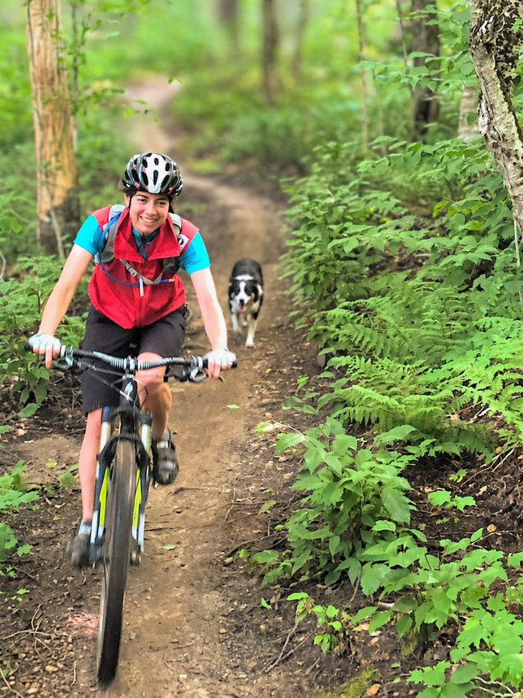 A girl riding a mountain bike on a dirt trail in a lush green forest, with a black and white dog following behind.