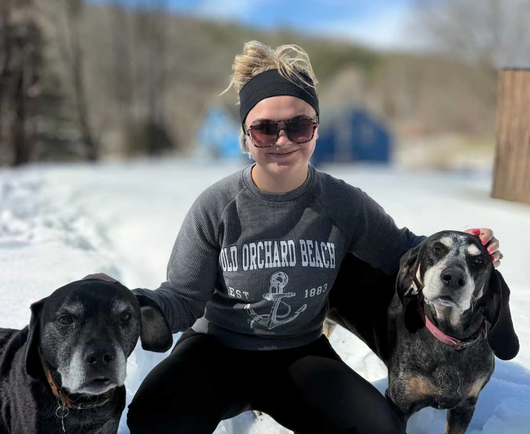 A woman wearing sunglasses, a black headband, and a gray sweatshirt with 'Old Orchard Beach' on it, smiling in the snow, with two dogs, one black with gray on its face, and a black and gray hound, posing outside on a snowy day.