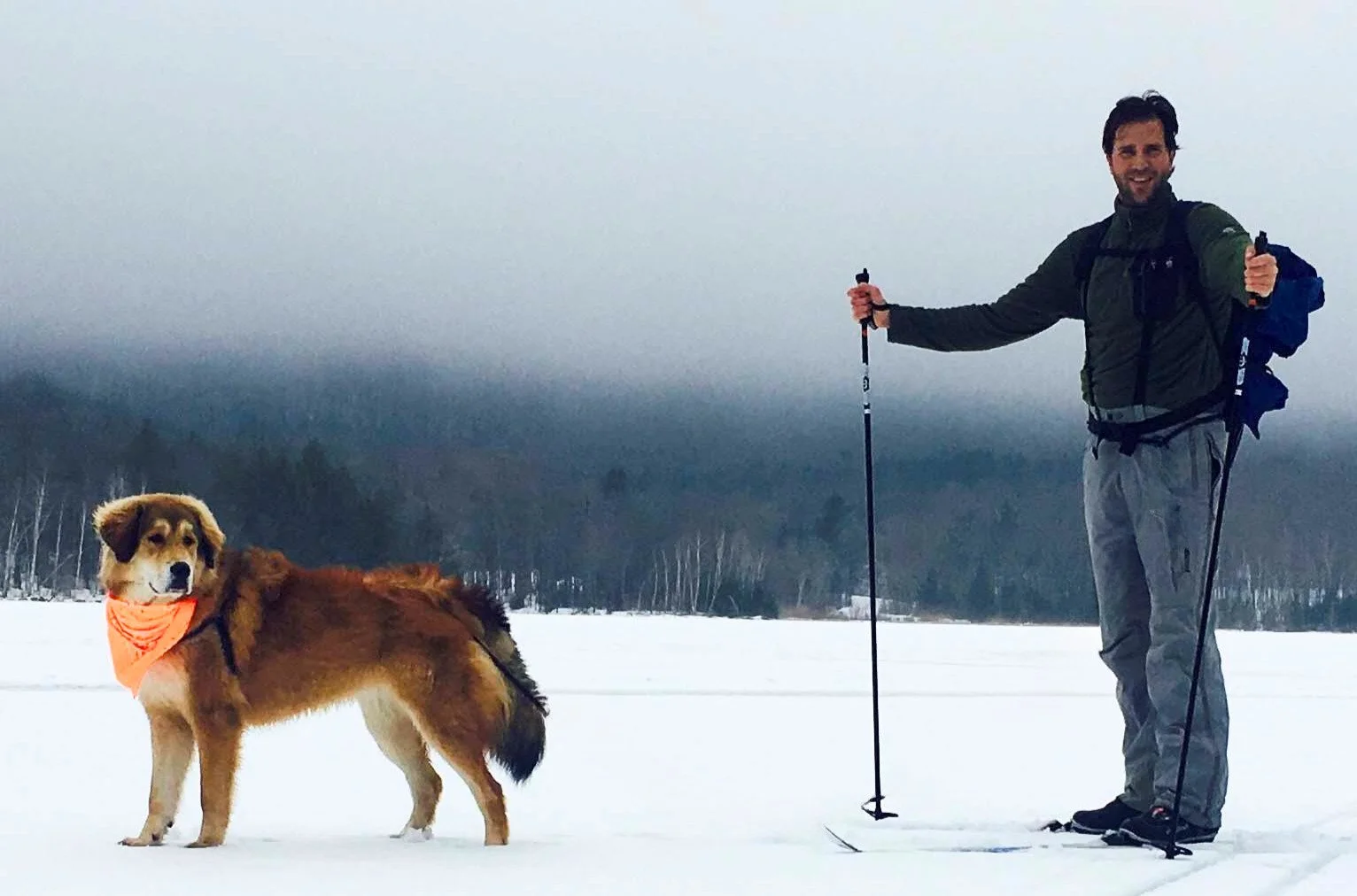 A man with a backpack and hiking poles standing on snow with a dog wearing an orange bandana, in a snowy landscape with a forested background.