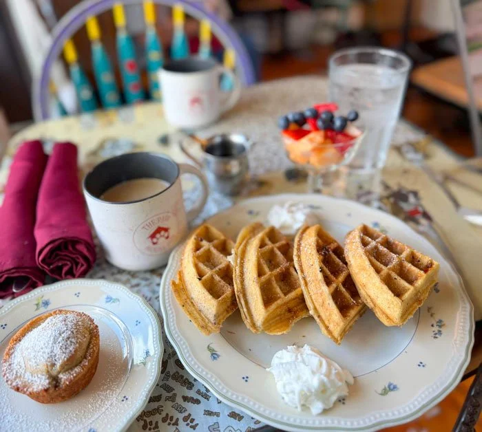A breakfast table with waffles, a muffin, fruit parfait and coffee in a mug stamped with The Paw House Inn