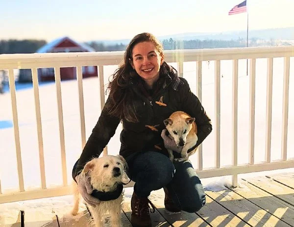 A woman is kneeling outdoors on a snowy deck, smiling, with two dogs, one on each side. The woman is holding a dog in her arms. An American flag is in the background.