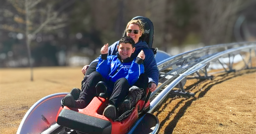 A woman and a young boy riding a roller coaster at an outdoor amusement park, smiling and enjoying the ride on a sunny day.