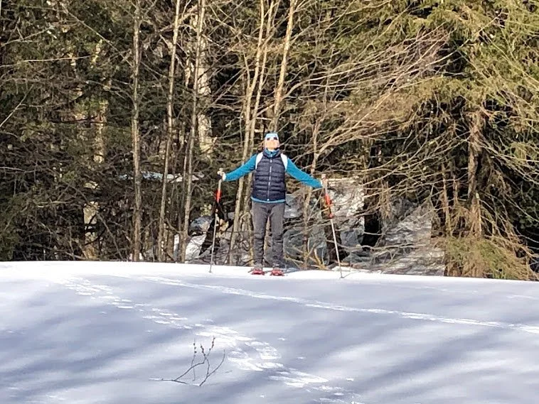 Person standing with ski poles in a snowy forested area, wearing a mask, sunglasses, and winter clothing.