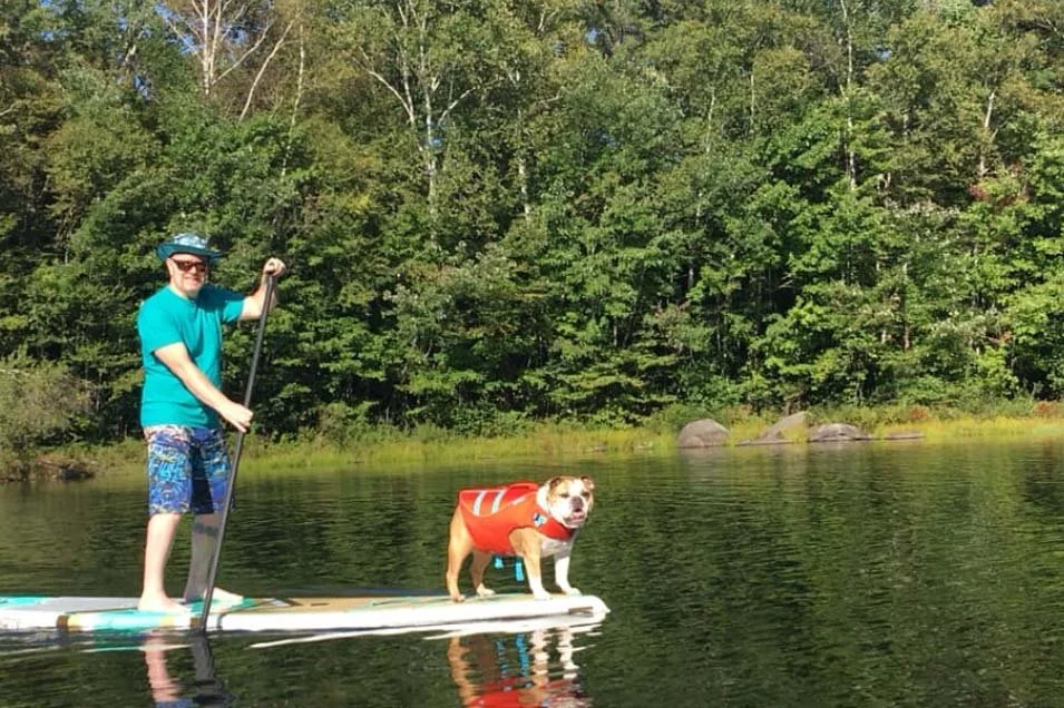 A man and his dog paddleboarding on a lake. The man is wearing a blue shirt, shorts, and a wide-brimmed hat, holding a paddle. The dog is standing on the paddleboard, wearing an orange life jacket.