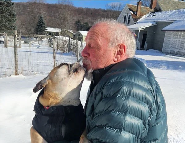 An elderly man and a dog sharing a kiss outside in a snow-covered yard, with houses and trees in the background.