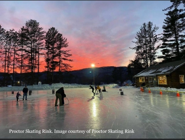 People ice skating on an outdoor rink at sunset, with trees and a wooden building in the background and orange cones on the ice.
