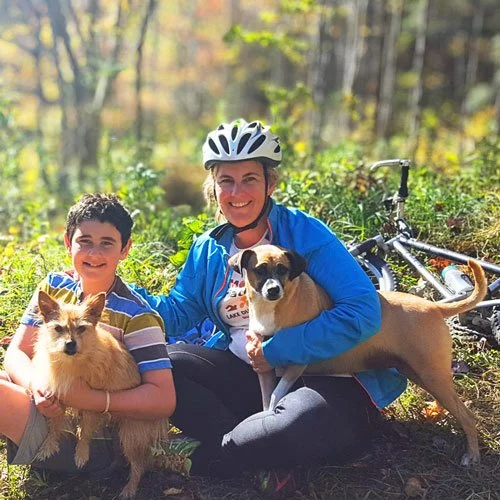 A woman and a young boy resting on a bike trail, holding two dogs, with a mountain bike lying on the ground nearby.