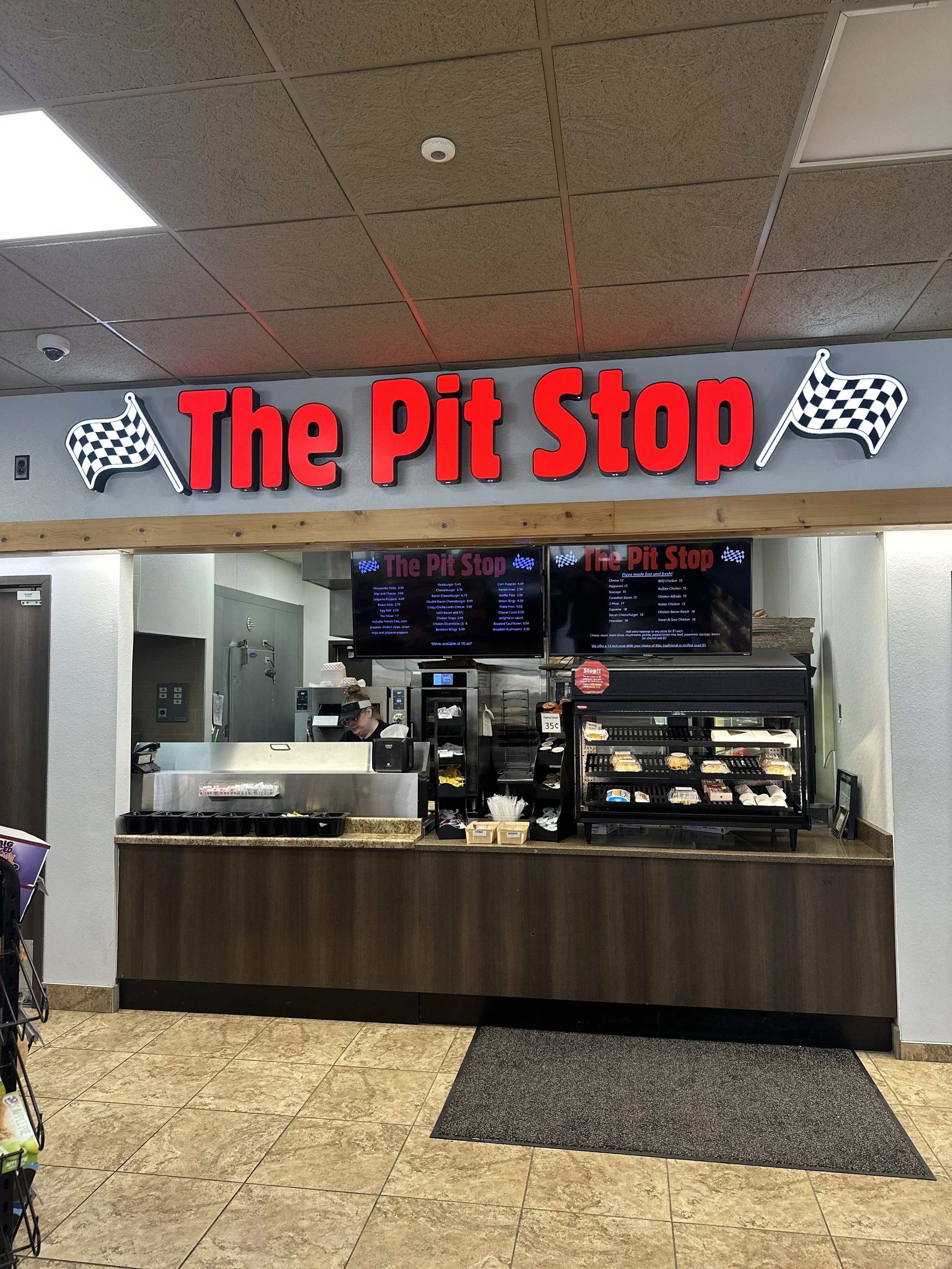 A fast food counter with a sign that reads 'The Pit Stop' in red letters, with checkered racing flags on either side. The counter has a glass display case with food items, and a worker behind the counter wearing a hat and glasses. digital menu screens are on the wall above the counter.