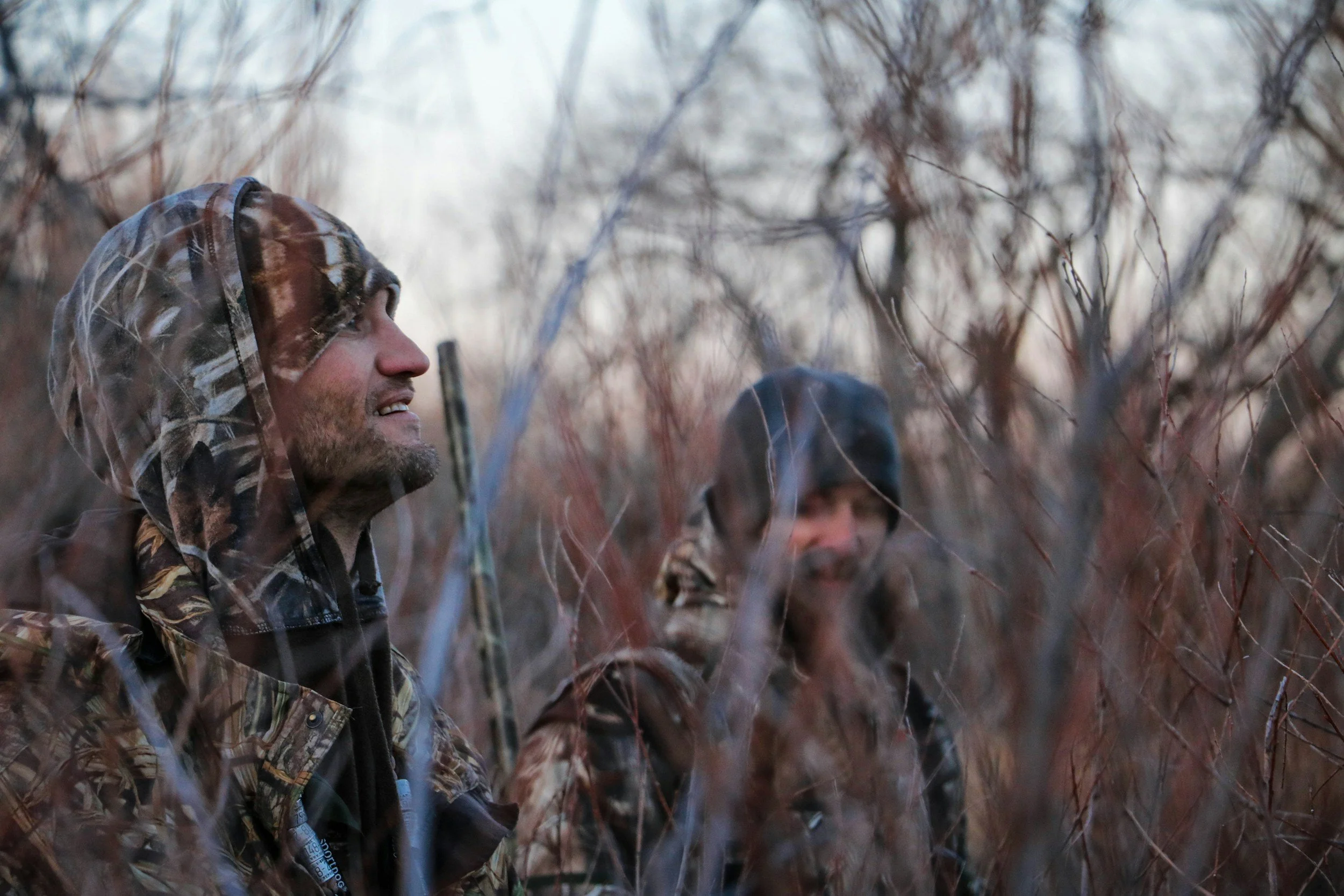 A man hunting with a camouflage hooded jacket smiling while standing outdoors among leafless trees and bushes.