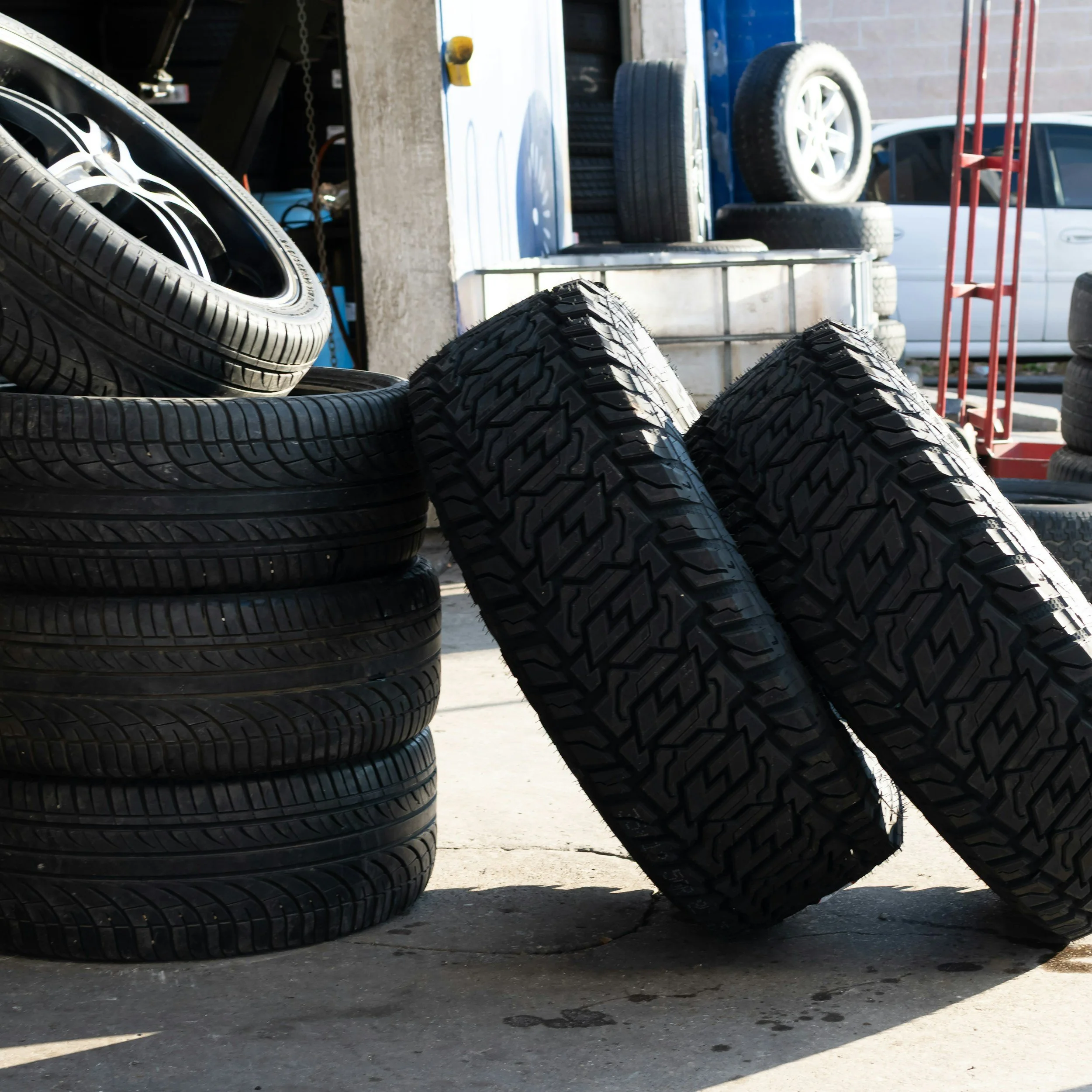 Stack of black tires with different tread patterns placed outdoors near a building with a truck in the background.