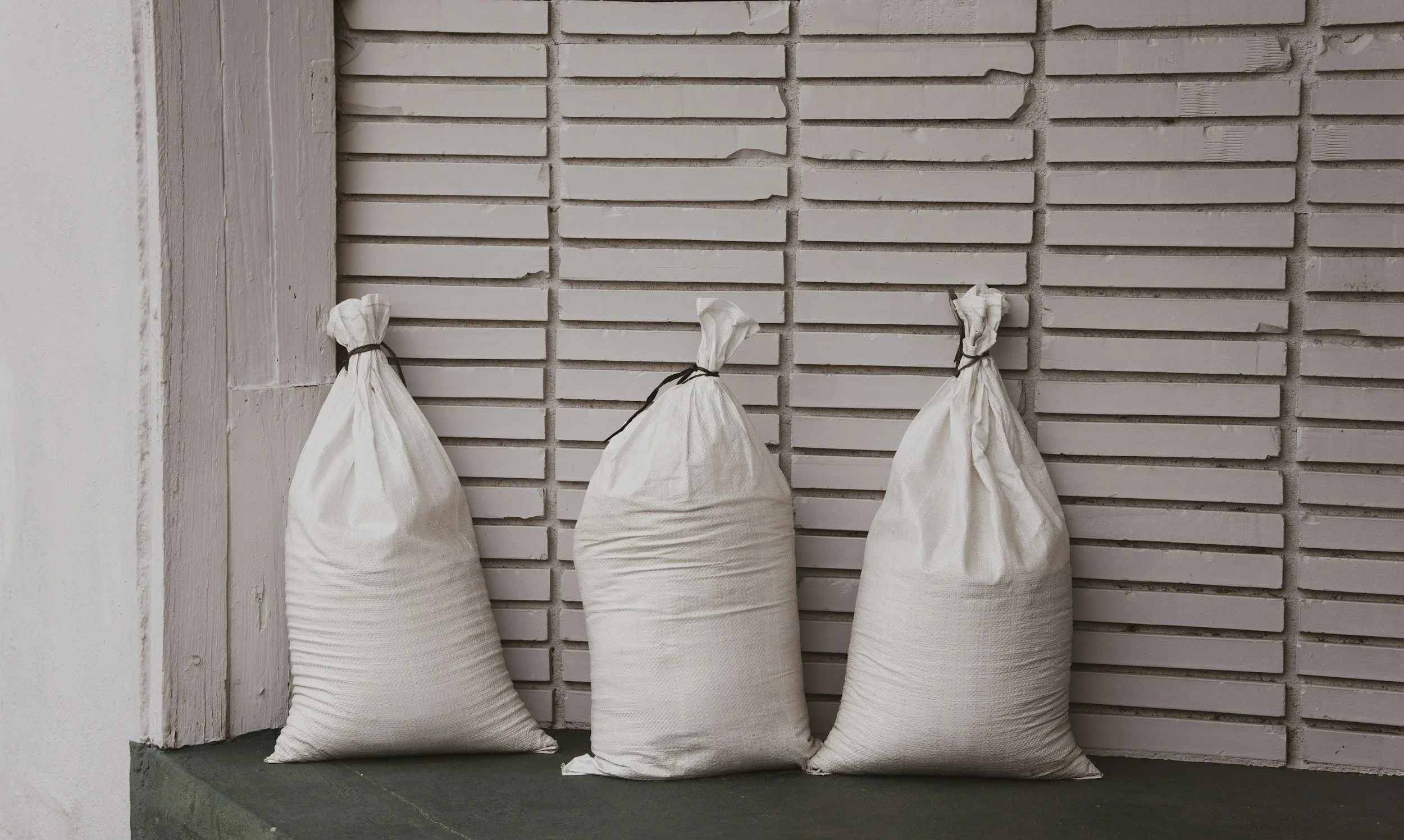 Four white cloth bags tied with black string, leaning against a white brick wall on a green concrete ledge.
