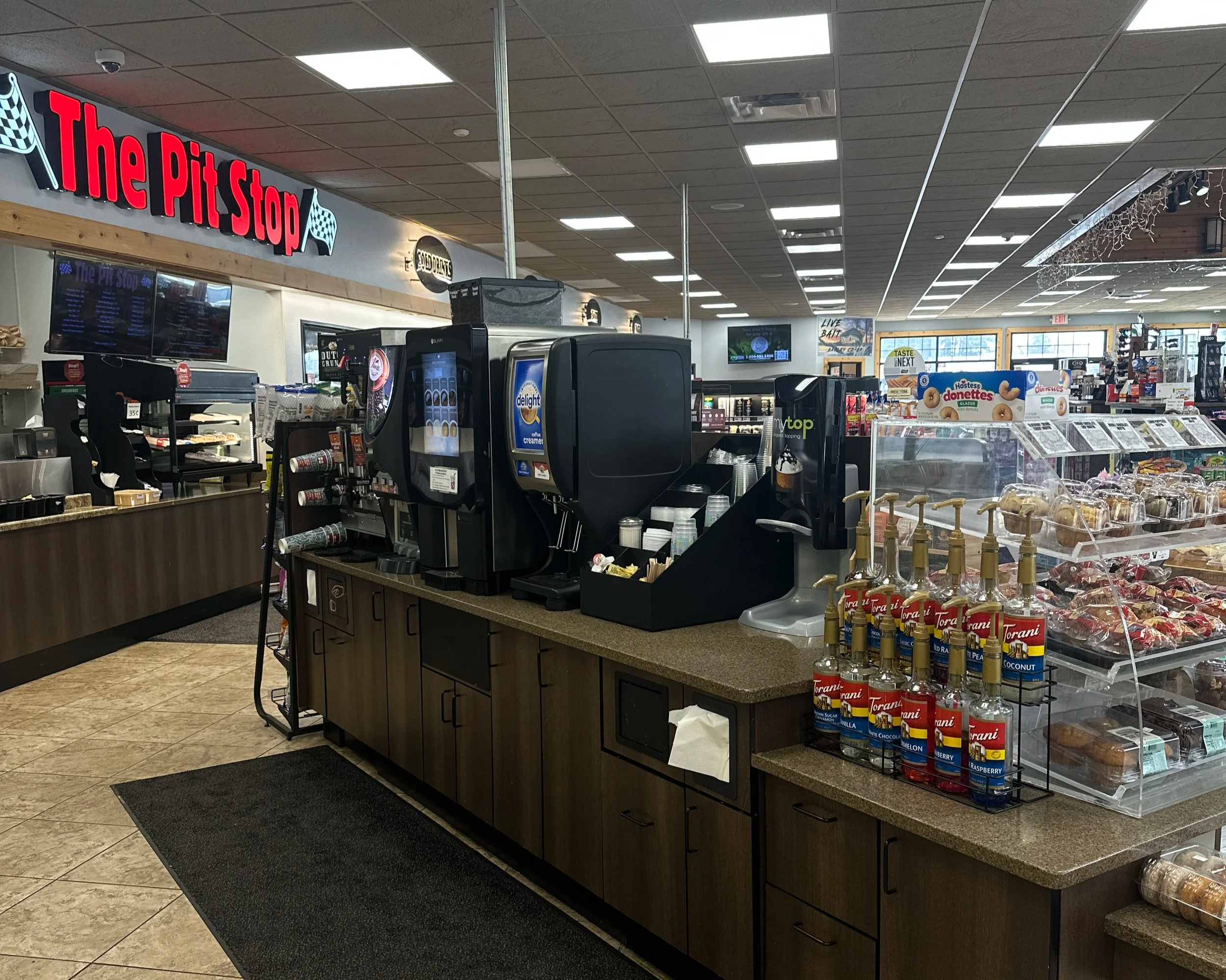 Interior of a convenience store or gas station with a counter and snack items, featuring a bright sign that reads 'The Pit Stop' with checkered flags, and various snack and beverage displays.