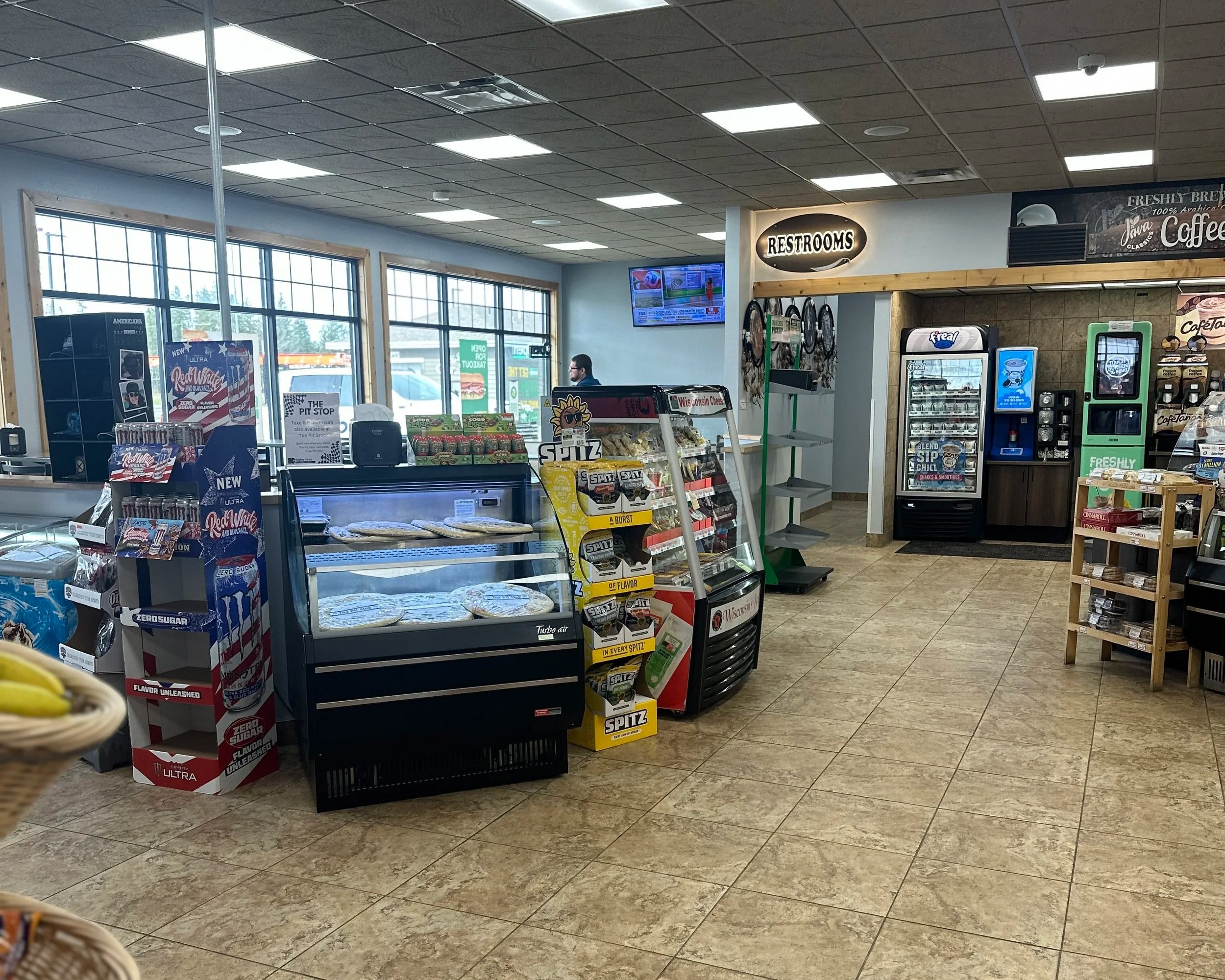 Inside a convenience store with snack displays, a checkout counter, and beverage vending machines near large front windows. A digital TV screen displays news and weather.