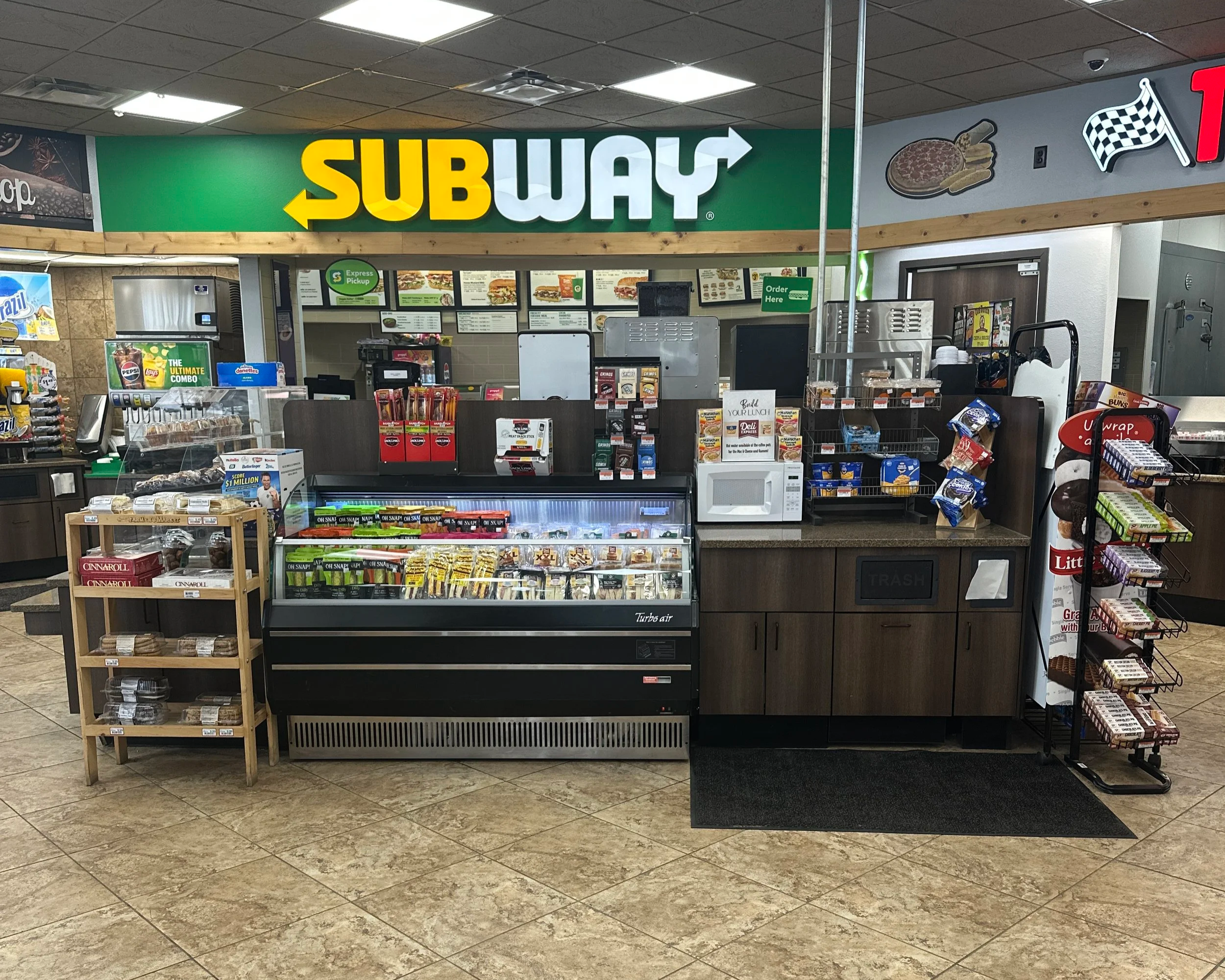 Interior of a Subway restaurant showing a menu board, a counter with various food items and snacks, and a display case with baked goods, with flooring and ceiling tiles.