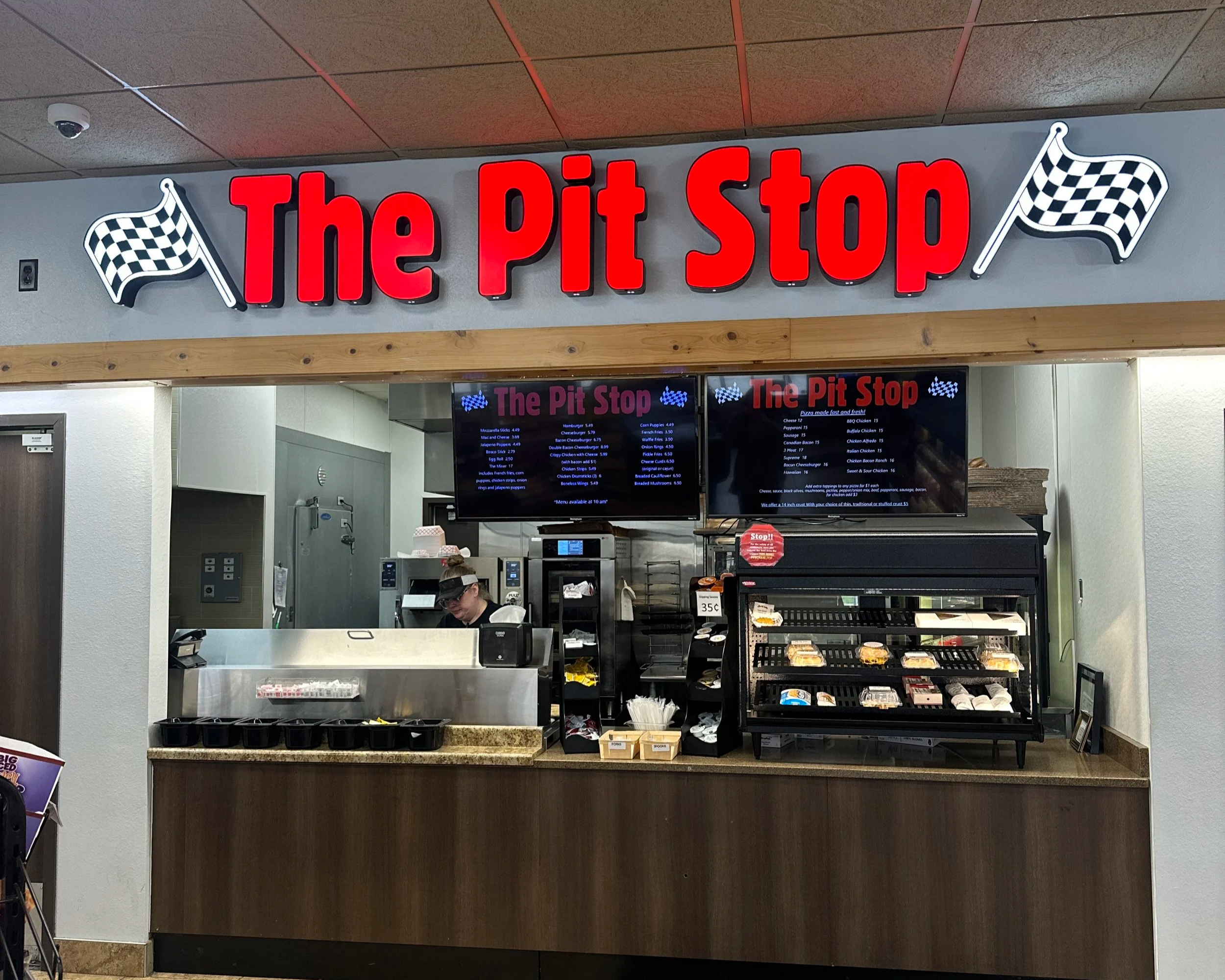 Sign for The Pit Stop with red lettering and checkered flags on a gray background, located above a food counter at a convenience store