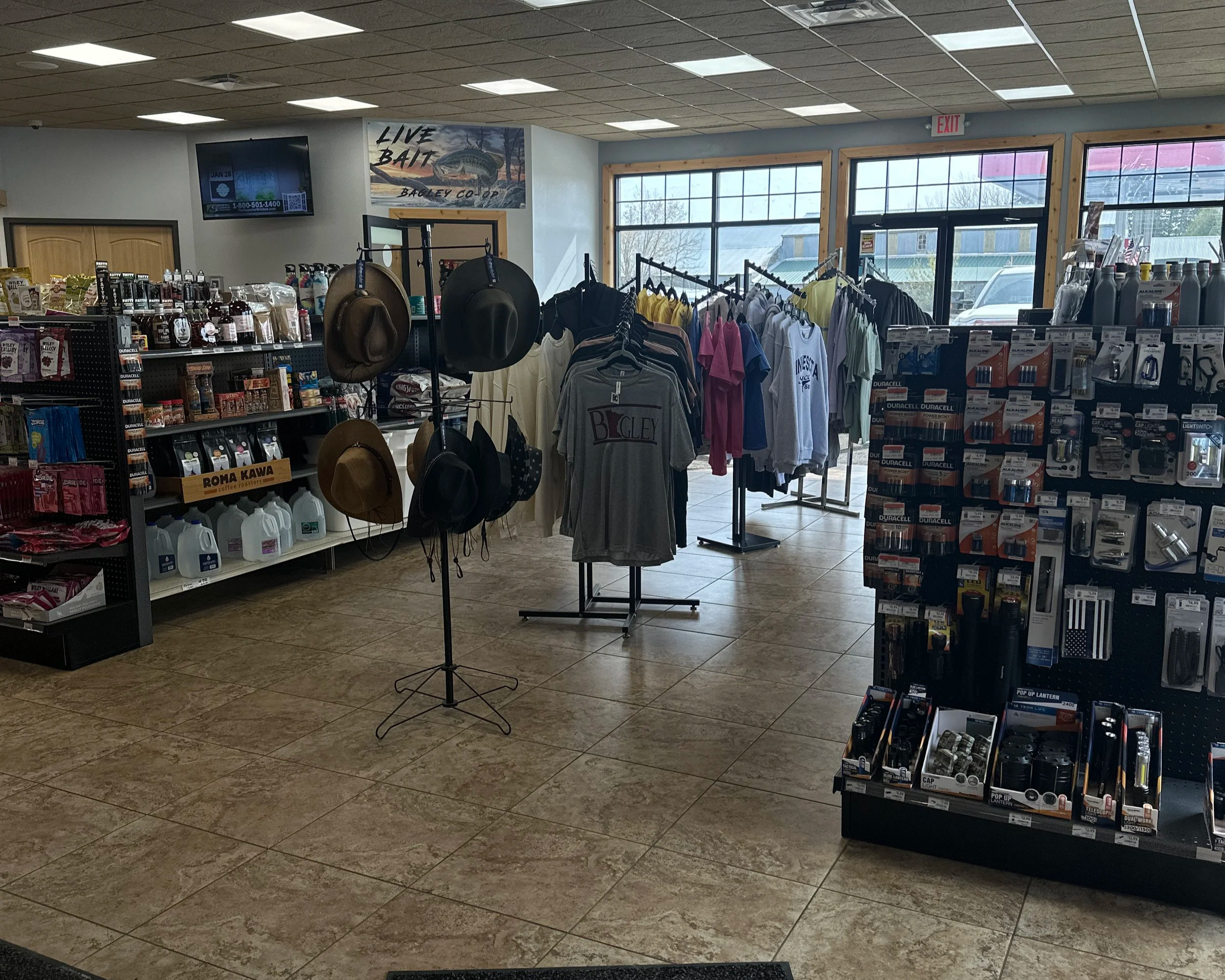 Interior of a convenience store with racks of hardware supplies, clothing, hats, and automotive items. Large windows let in natural light, and a sign reads 'Live Bait' with a fish illustration.