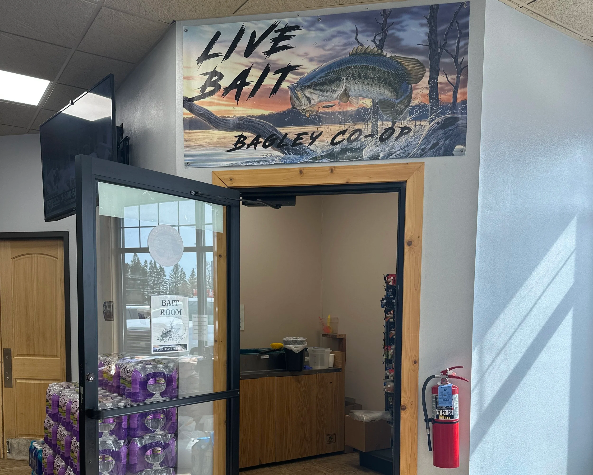 Poster with a fish jumping out of water at sunset, with the words "LIVE BAIT" and "BAGLEY CO-OP" on it. Inside the store, there are shelves with bottled water, a fire extinguisher, and a door with a sign that reads "BAIT ROOM."