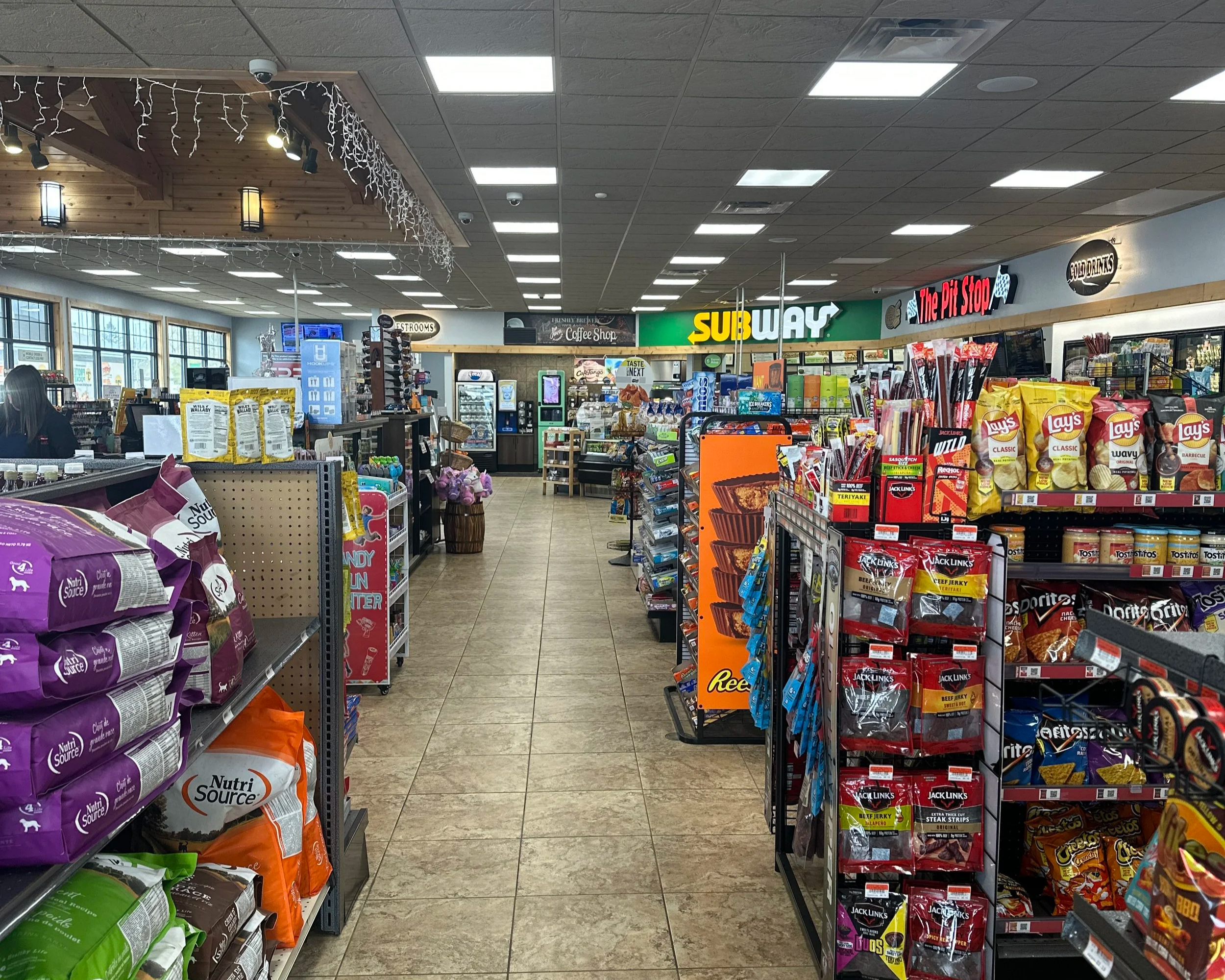 View of an aisle in a convenience store with shelves on both sides stocked with snacks, chips, and other food items. The ceiling has recessed lighting, and there are signs for Subway, The Pit Stop, and other sections visible.