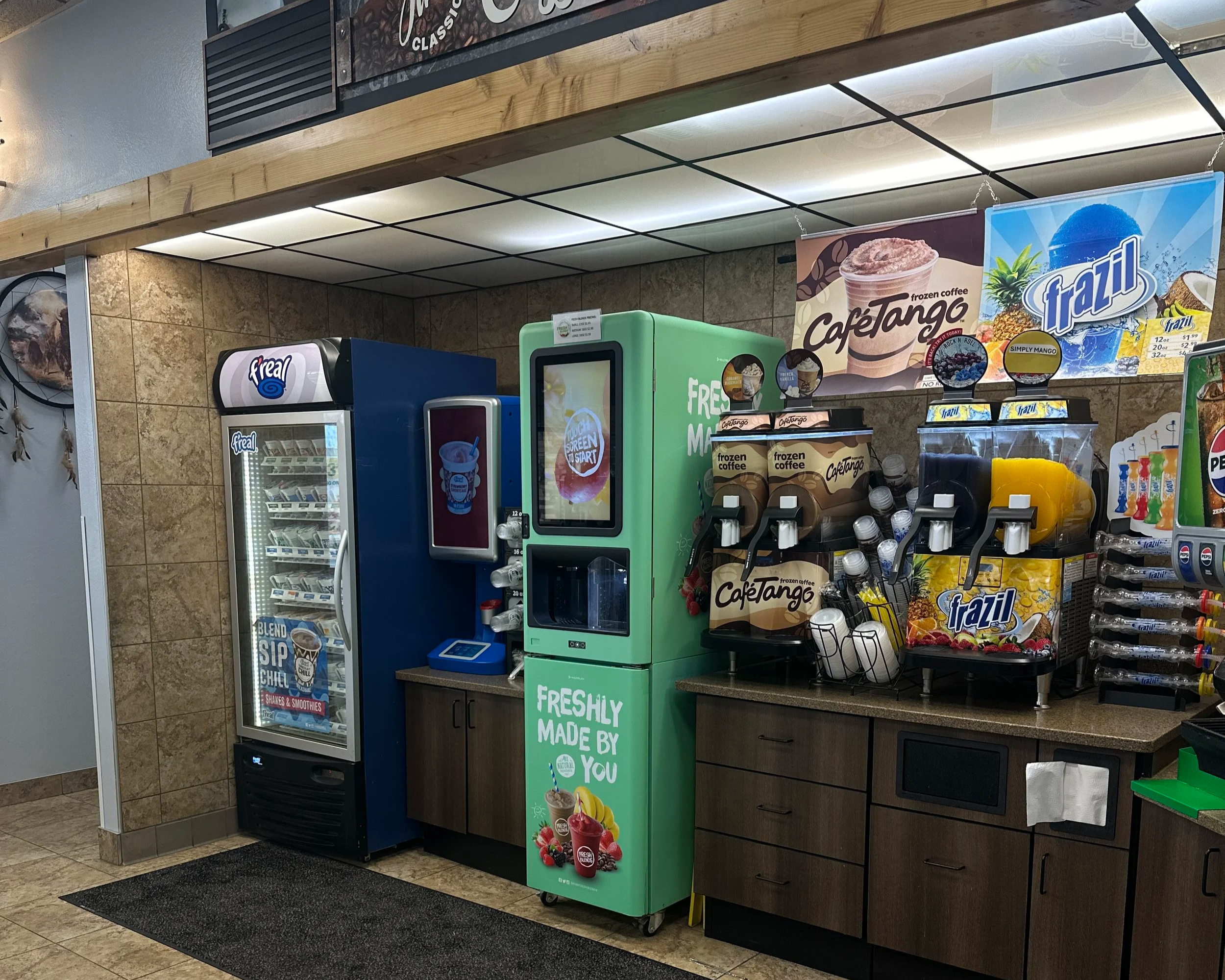 Inside a convenience store, a frozen beverage station with frozen coffee drinks from CafeTango, next to smoothie options. A vending machine is on the left, and various snacks and drink accessories are displayed on shelves. The ceiling has bright fluo